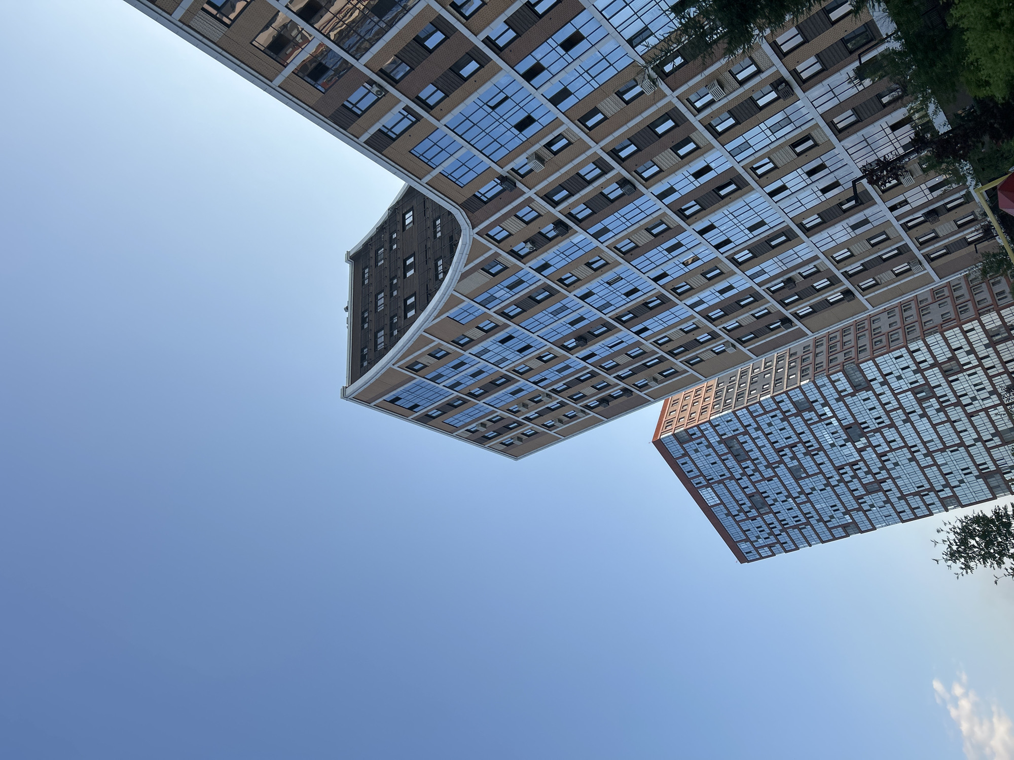 The media file shows an upward-looking perspective of modern apartment buildings against a clear blue sky. The scene is dominated by the architecture, with two distinct buildings prominently featured.
The building on the left is large and has a curved facade. Its exterior is a mix of light brown brick and dark sections, with a grid pattern of windows. Many of these windows are reflective, mirroring the sky. Some windows are smaller and square, while others are longer and rectangular. The building on the right is also tall and appears to be made of brick with a facade covered in numerous windows. These windows are mostly rectangular and reflect the sky, giving the building a shimmering appearance.
The perspective suggests the photo was taken from the ground level or a lower vantage point. The angle emphasizes the height and scale of the buildings. There are some trees visible on the right side of the frame, suggesting an urban or suburban setting with some greenery.
The time of day appears to be daytime, judging by the bright sky and clear visibility. The weather seems to be fair and sunny. There is no visible text in the image. The overall impression is one of modern urban development and architectural design.