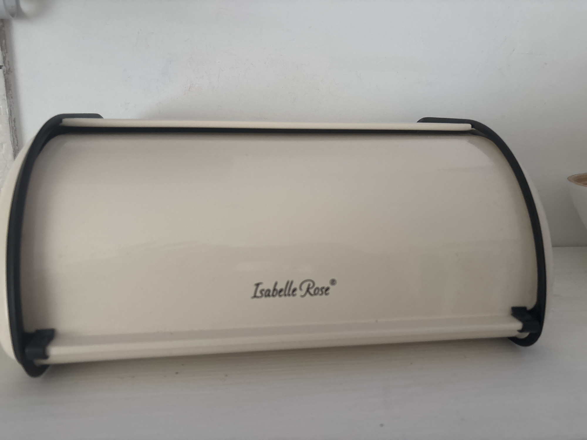 A close-up photograph shows a breadbox with the brand name "Isabelle Rose" in cursive font, set against a white wall and surface. The breadbox is primarily cream-colored with black trim. A small portion of a white bowl is visible on the right edge. The image lacks any specific clues linking it to Saint Petersburg, Russia. The setting appears to be indoors, possibly a kitchen or pantry area.