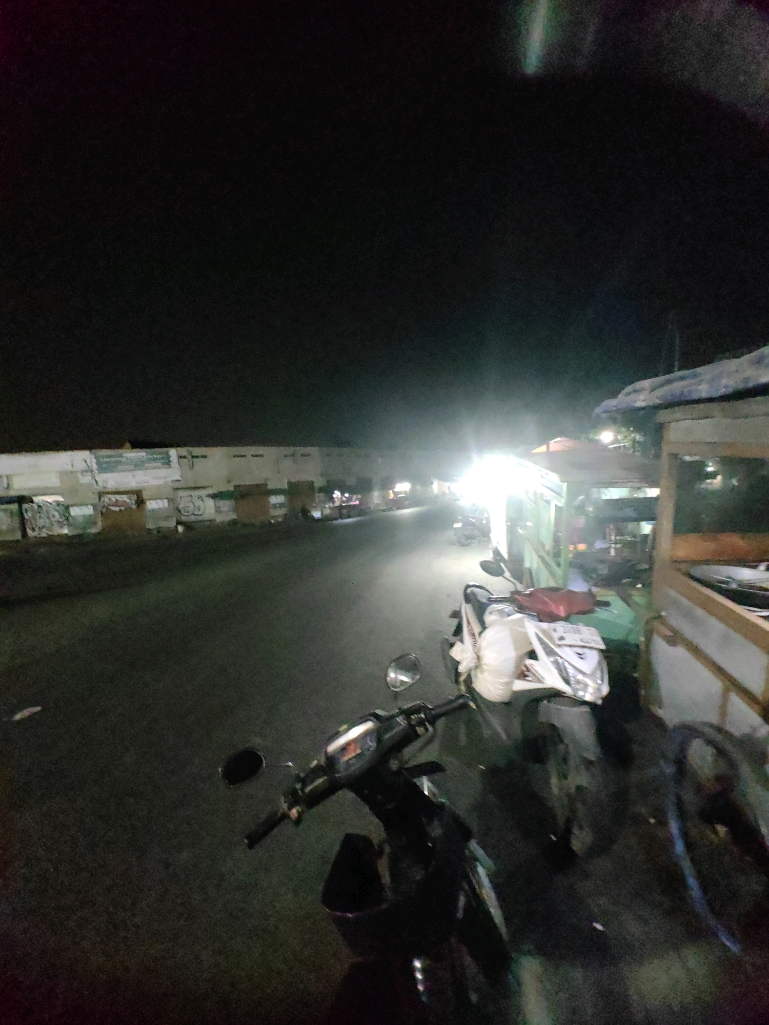 In Keboansikep, Indonesia, a nighttime scene unfolds on a paved road. The image is dominated by the darkness of night, with artificial lights creating strong contrasts. In the foreground, two motorcycles are parked. The motorcycle closer to the viewer is dark-colored, with a black seat and handlebars. Further back, a white scooter with red accents is visible, carrying a white plastic bag on its front. To the right of the motorcycles, a wooden food stall or cart is partially visible, with a light-colored tarp overhead. The background shows a row of buildings or structures lining the road, some of which appear to have graffiti or posters. The road itself is dimly lit, with a bright light source emanating from further down the street, casting long shadows. The overall impression is one of a quiet, urban setting at night, possibly near a marketplace or a commercial area.
