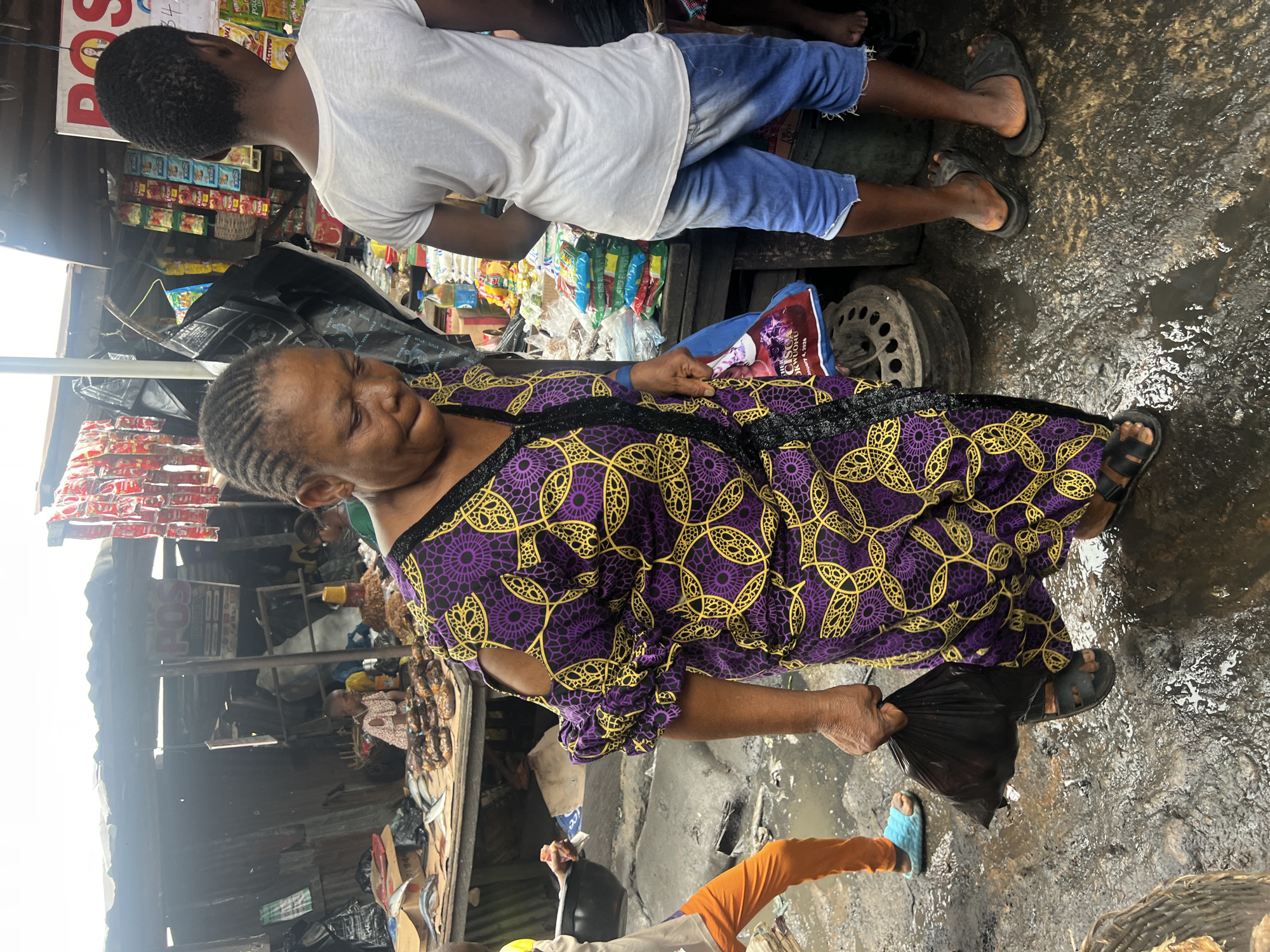 The image depicts a busy marketplace scene in Nkpor, Nigeria. A woman in a purple and yellow patterned dress is walking and carrying a black plastic bag in her left hand. She is wearing black sandals. To her right, a person in a white t-shirt and denim shorts is standing. In the background, there are stalls displaying various goods, including food items like fish and packaged products. A young child is visible sitting near one of the stalls. The ground is wet and appears to be muddy, suggesting recent rain or a damp environment. The overall atmosphere is that of a bustling local market.