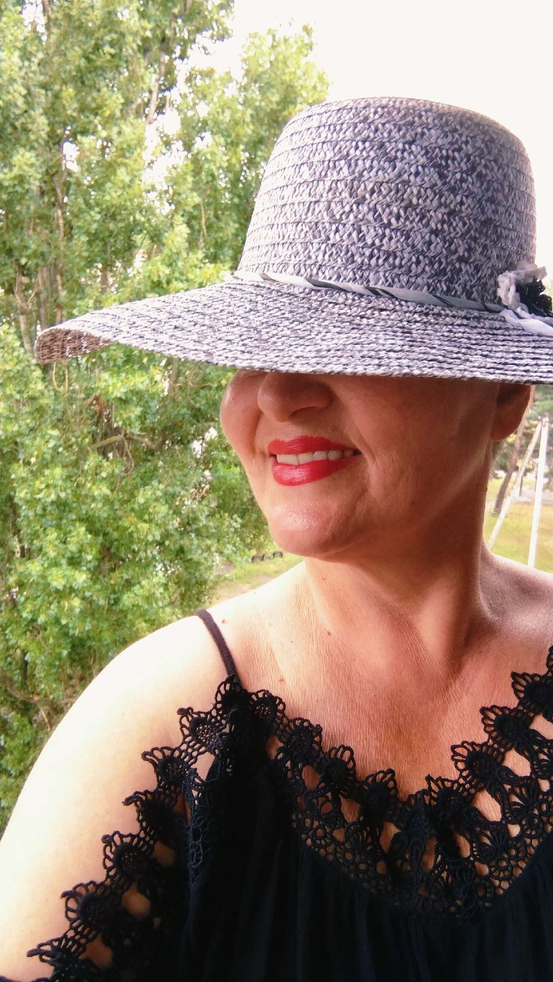A woman wearing a wide-brimmed straw hat with a gray ribbon and a black lace trim dress is depicted in an outdoor, possibly residential setting. The woman has bright red lipstick and is smiling. The background consists of lush green foliage, suggesting a natural or garden environment. The image was taken in the city of Horishni Plavni, Ukraine, likely on a balcony or porch given the visible railing and railing support in the background.
