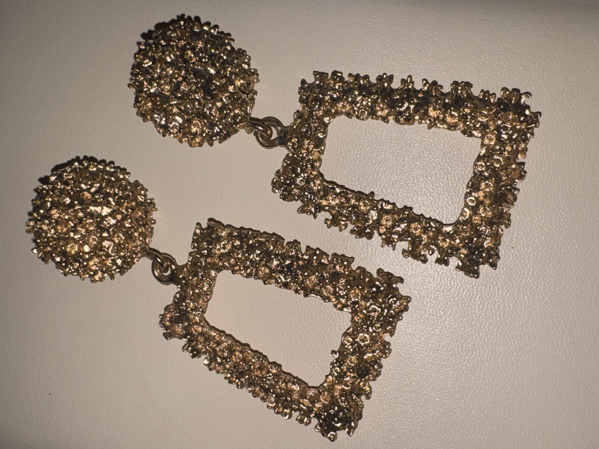 The image shows a pair of gold-colored earrings against a neutral, light-colored background, likely a surface. Each earring consists of a circular, textured stud connected by a small gold ring to a rectangular, hollow, textured pendant. The texture of the earrings appears rough and faceted, giving them a somewhat raw or organic look, and they catch the light, creating highlights and shadows. The earrings are laid out side-by-side, with the top earring positioned slightly above and to the right of the bottom earring. The lighting suggests an indoor setting, possibly a studio or a home, with a single light source casting shadows. There is no text visible in the image. The context provided, "Яромінскі сельскі Савет, Belarus," indicates a rural area in Belarus, but this geographical information is not visually discernible from the image itself. The scene is static, focusing solely on the display of the jewelry.