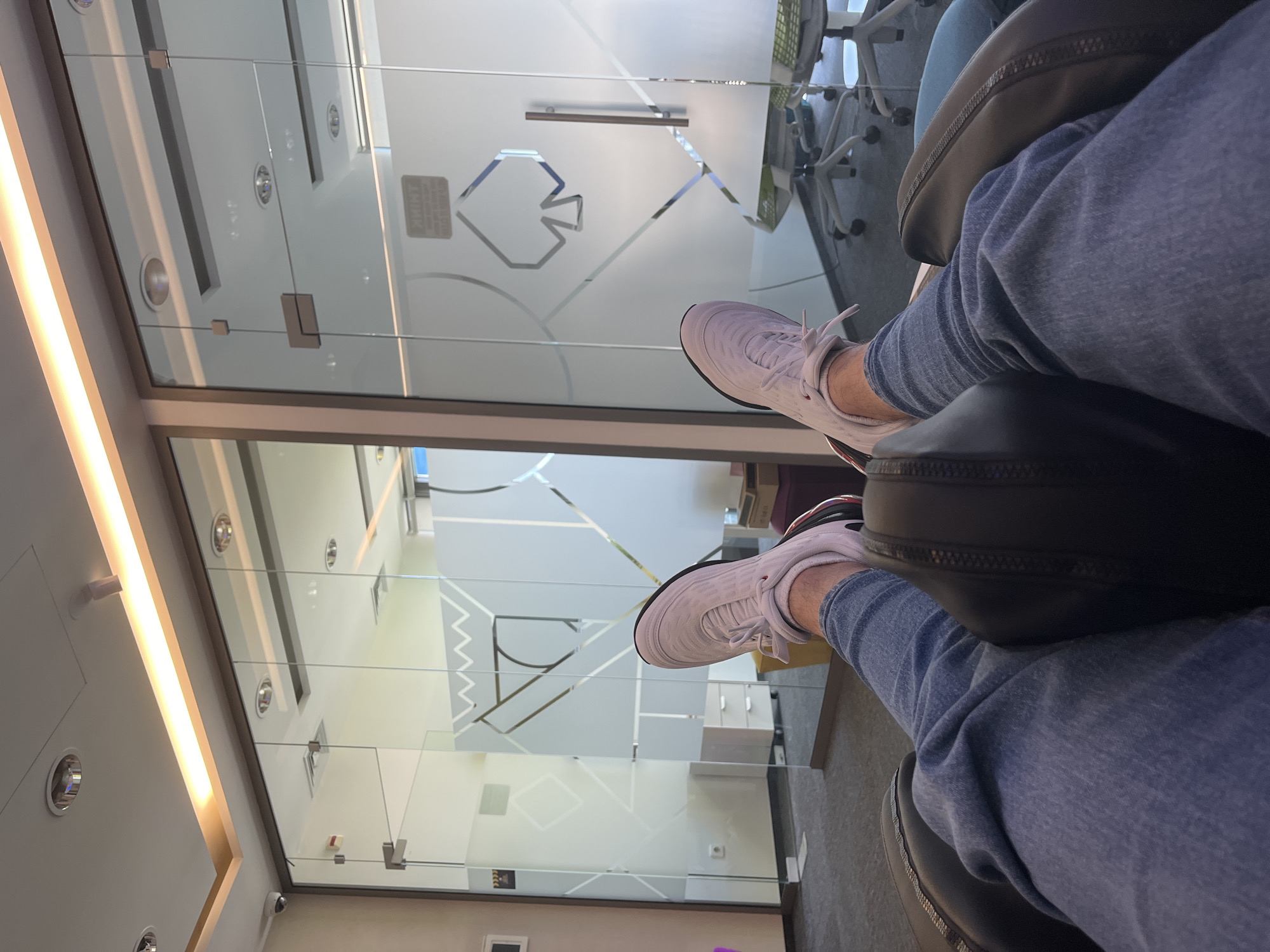 The image depicts a seated individual with their legs crossed, wearing light blue jeans and white sneakers. The setting appears to be an office environment, evident from the glass-walled office spaces behind the individual, featuring patterned glass designs. The lighting fixtures and interior layout suggest a modern office building. There is no identifiable information to confirm the location in Unknown, Unknown.