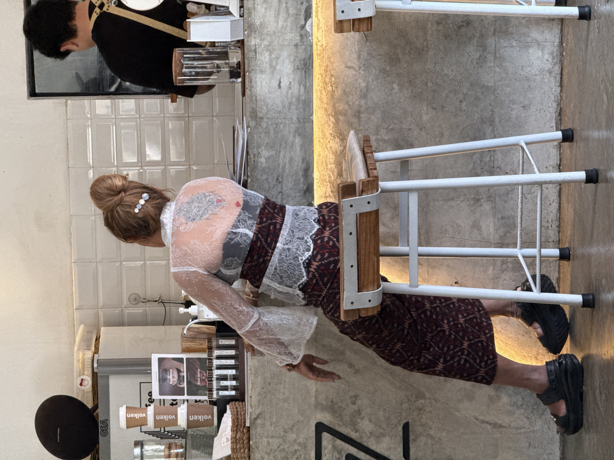 In a bright, modern cafe in Kuta, Indonesia, a woman with auburn hair styled in a high bun sits facing away from the camera, resting her left elbow on a concrete counter. She wears a white lace top with flared sleeves over a black undergarment and a dark patterned skirt. Her back is adorned with a large, intricate tattoo. On the counter in front of her are several small glass vials with labels, a woven basket, and stacked coffee cups. To her right, a barista with dark hair and a black shirt with a tan apron stands behind the counter, partially visible. The cafe features white tiled walls and a polished concrete floor, with modern stools and a counter illuminated by warm yellow lighting.