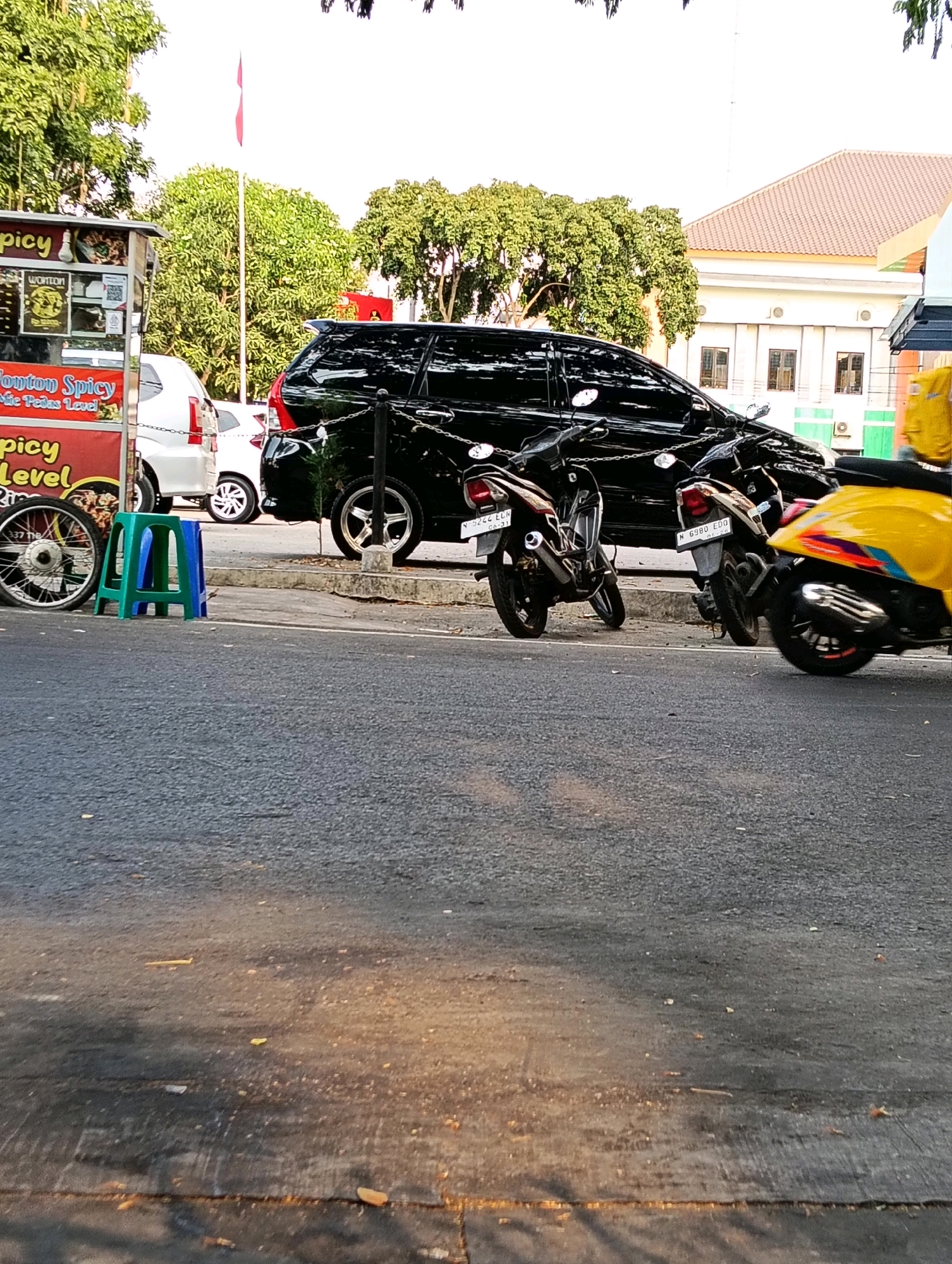 The image depicts a street scene in Karanganyar, Indonesia, with a focus on a food cart on the left, a black SUV in the middle, and several motorcycles and a yellow scooter on the right. The food cart displays menus with text including "Spicy" and "Level." A red and white striped tarp is visible on the cart. A white car is parked behind the food cart. The black SUV is parked parallel to the street, with its rear facing the viewer. Several motorcycles are parked behind the SUV, and a yellow scooter is to the far right, partially obscuring a black motorcycle. A white building with a tiled roof and windows is visible in the background, along with trees and a flagpole with a red and white flag. The street surface is asphalt, with some debris and shadows cast across it. The sky is overcast.