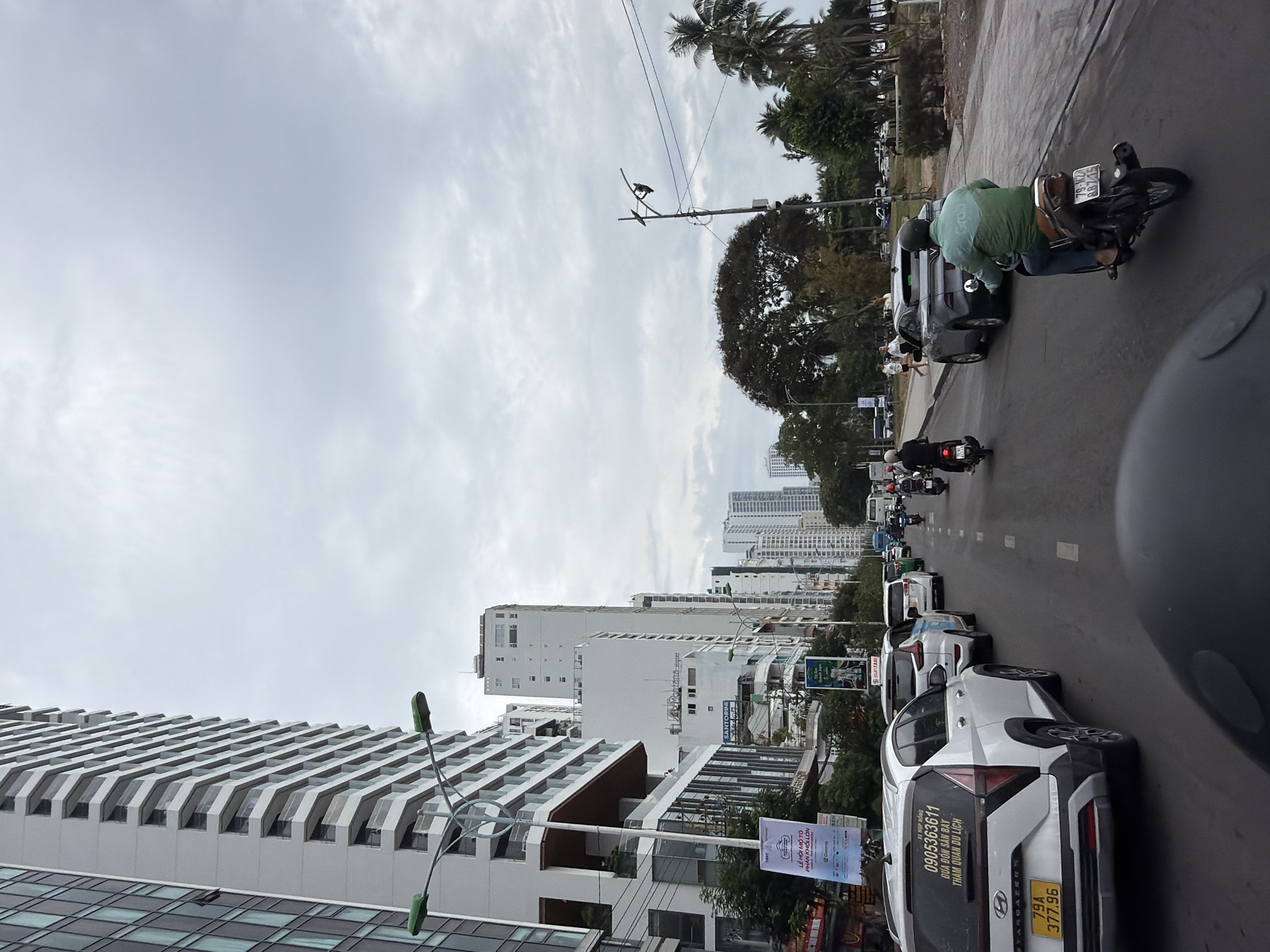 The image depicts a street scene in what appears to be Nha Trang, Vietnam. The shot is taken from a street-level perspective, possibly from within a vehicle or on the street itself.
Dominating the foreground is a line of parked cars on the right side of the road. A white SUV with a license plate "79A 377.96" and a taxi advertisement on the back is prominently visible. A banner advertising a motorbike festival and a local transportation service is also visible.
In the background, there's a moderate flow of motorcycle traffic heading away from the camera, interspersed with cars. Tall buildings, typical of a city skyline, line the horizon. The sky is overcast, suggesting a cloudy day.
A motorcyclist wearing a green shirt is interacting with a dark-colored vehicle. Palm trees and other greenery are visible in the background, adding to the urban landscape.