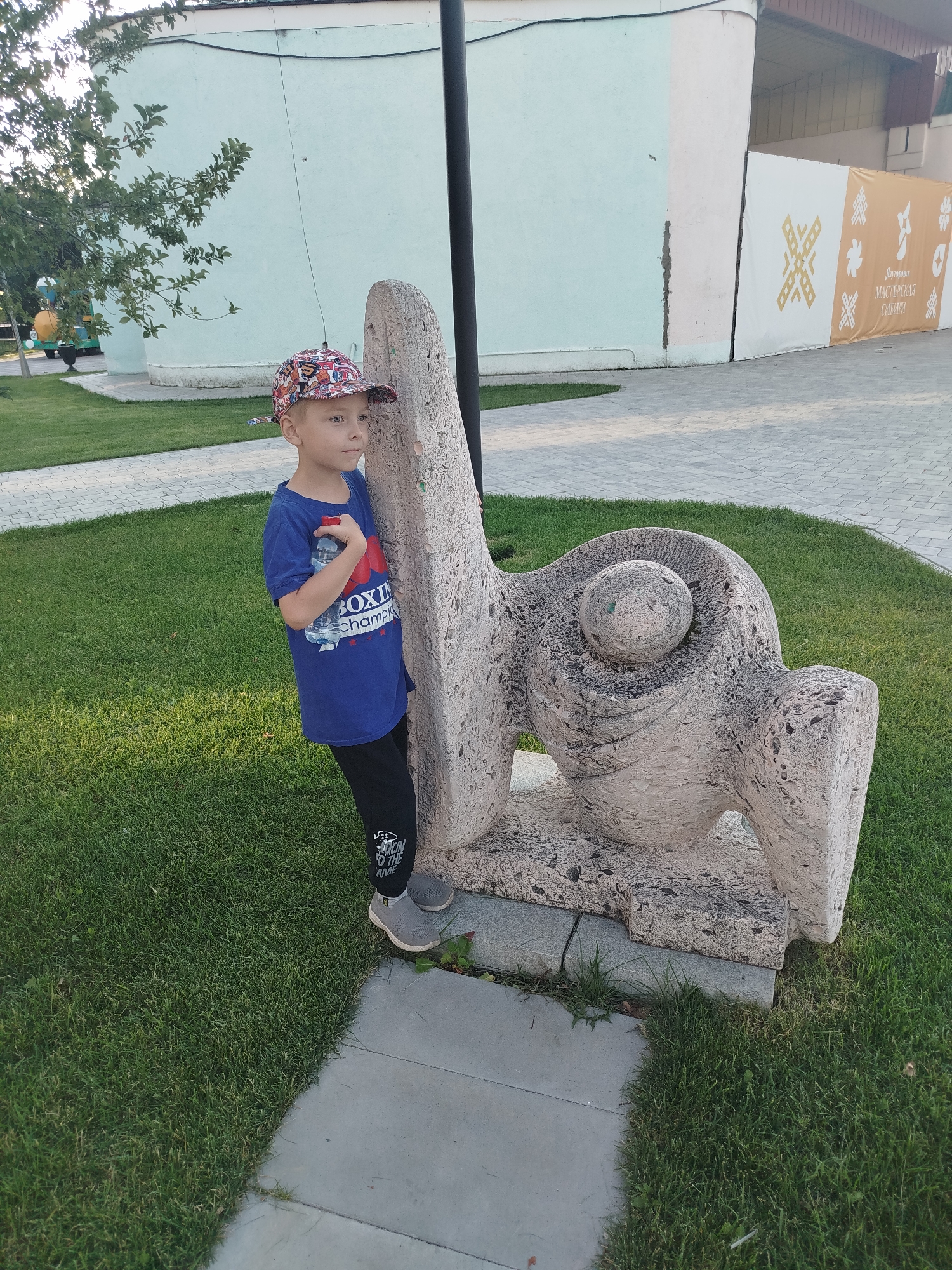 A young boy wearing a blue t-shirt, black pants, a patterned baseball cap, and gray sneakers stands to the left of a concrete sculpture in a park. The boy is holding a clear water bottle. The sculpture appears to be abstract. In the background, a light blue building is visible, along with a fence with an orange banner. The park has a grassy area and a paved pathway. The scene is lit by natural sunlight, suggesting it is daytime. The location is identified as a park in the city of Yalutorovsk, Russia.