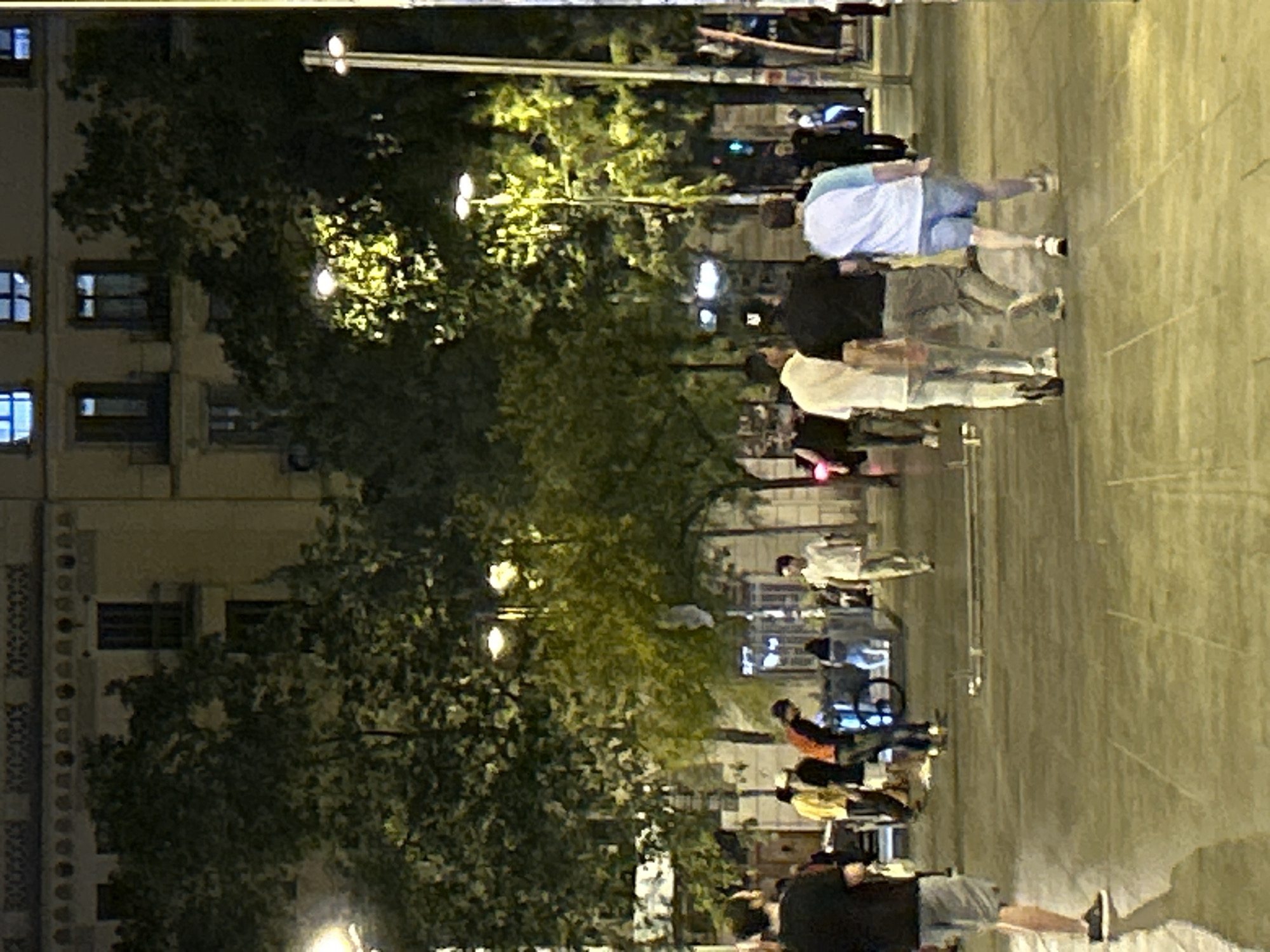 The image depicts a nighttime street scene in Barcelona, Spain, likely in a public square or pedestrian area. In the foreground, several individuals are walking. To the left, a person wearing a black t-shirt and grey shorts is walking away from the camera. Further into the scene, a group of three men are walking together; one in a white shirt and light-colored pants, another in a black t-shirt and dark pants, and the third in a white and light blue t-shirt with blue shorts. To the right of this group, another person in a light-colored shirt and pants is walking away. In the background, several trees with illuminated foliage are visible, interspersed with buildings featuring illuminated windows. Streetlights cast a yellowish glow on the scene. The pavement is paved with rectangular tiles. The overall atmosphere suggests a lively urban environment at night.