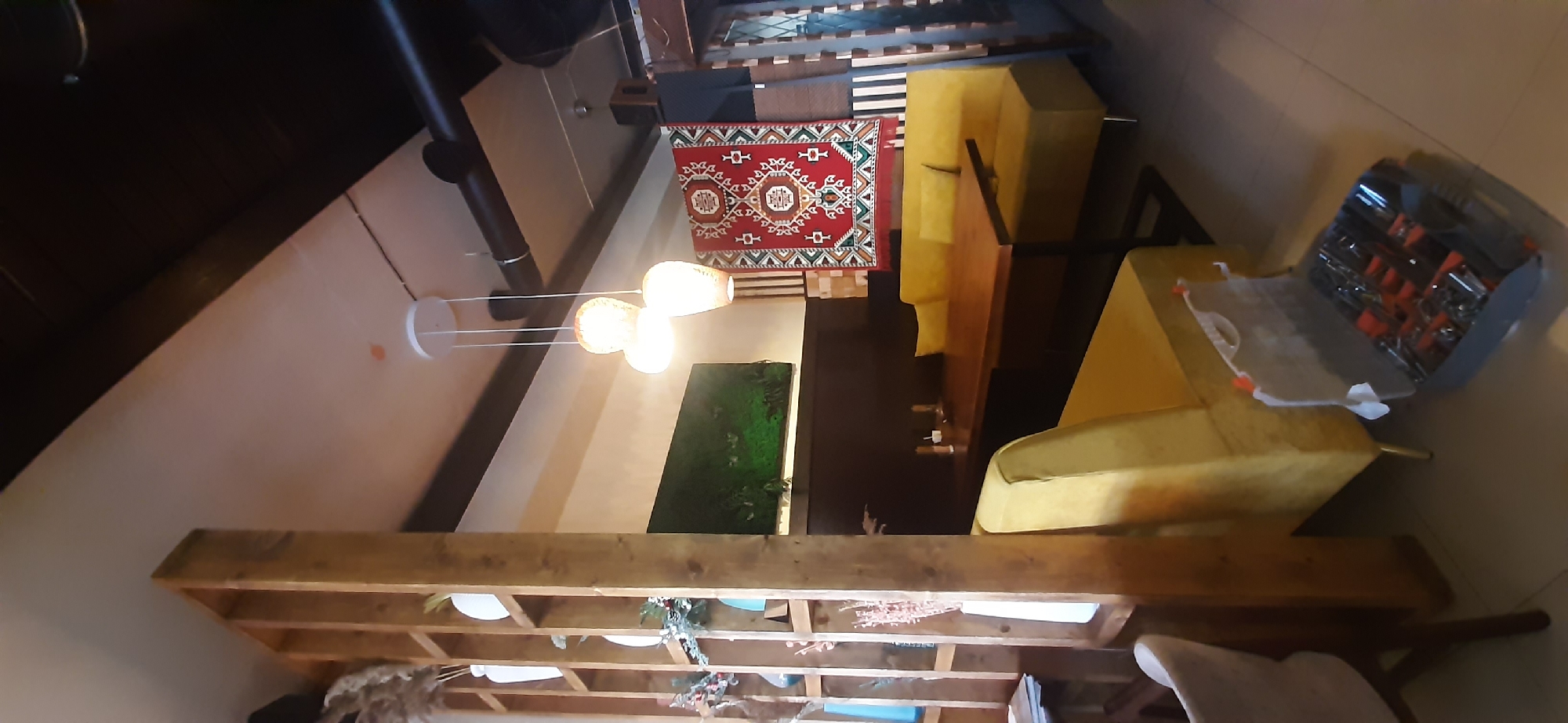 The image depicts an interior scene, likely a home or a commercial space, possibly a lounge area. The scene is shot from a slightly elevated perspective, with the camera tilted. In the foreground, a wooden bookshelf with several compartments is visible, displaying some decorative items and plants. Above the bookshelf, a modern chandelier with three spherical light fixtures hangs from the ceiling. To the left, a dark, cylindrical column, possibly a chimney or support beam, runs vertically.
In the middle ground, a red and white patterned rug is laid out on the floor. Behind the rug, there's a plush yellow sofa, with another yellow upholstered piece of furniture visible further to the right. A green moss-covered panel, possibly a piece of wall art or a terrarium, is mounted on the wall. Further to the right, a gray toolbox with various tools, predominantly orange and silver, is open on the yellow furniture.
The lighting suggests it could be daytime or artificial lighting is used, creating a warm ambiance. The overall setting appears to be indoors, with architectural elements like wooden beams and a sloping ceiling suggesting a contemporary or possibly a loft-style space. No people are visible in the image, and there is no discernible weather or time of day cues. No visible text is present. The location context provided is Yessentuki, Russia, which might suggest a Russian interior design style, although the current visual information does not strongly confirm this. The arrangement of furniture and decor indicates a designed space for relaxation or socializing.