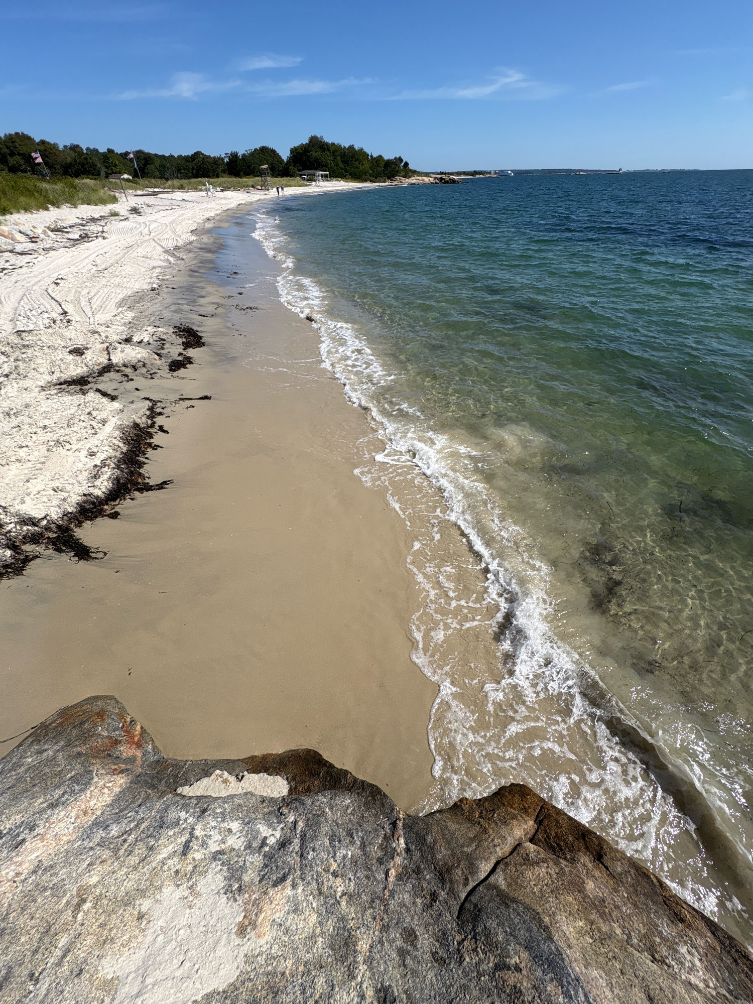 A bright, sunny day at a sandy beach is depicted in this image, located in Waterford, Connecticut, USA. The foreground is dominated by a large, grey and white rock. The beach itself is composed of fine, light-colored sand, with some patches of dark seaweed along the tideline. Gentle waves are breaking on the shore, with clear, green water and visible sandy bottom. In the mid-ground, a line of green trees and vegetation borders the beach, with a few buildings and structures visible among them, including what appears to be a lifeguard station and a small booth. Several people can be seen walking along the shoreline and in the water. The sky is a clear, vibrant blue with a few wispy clouds.