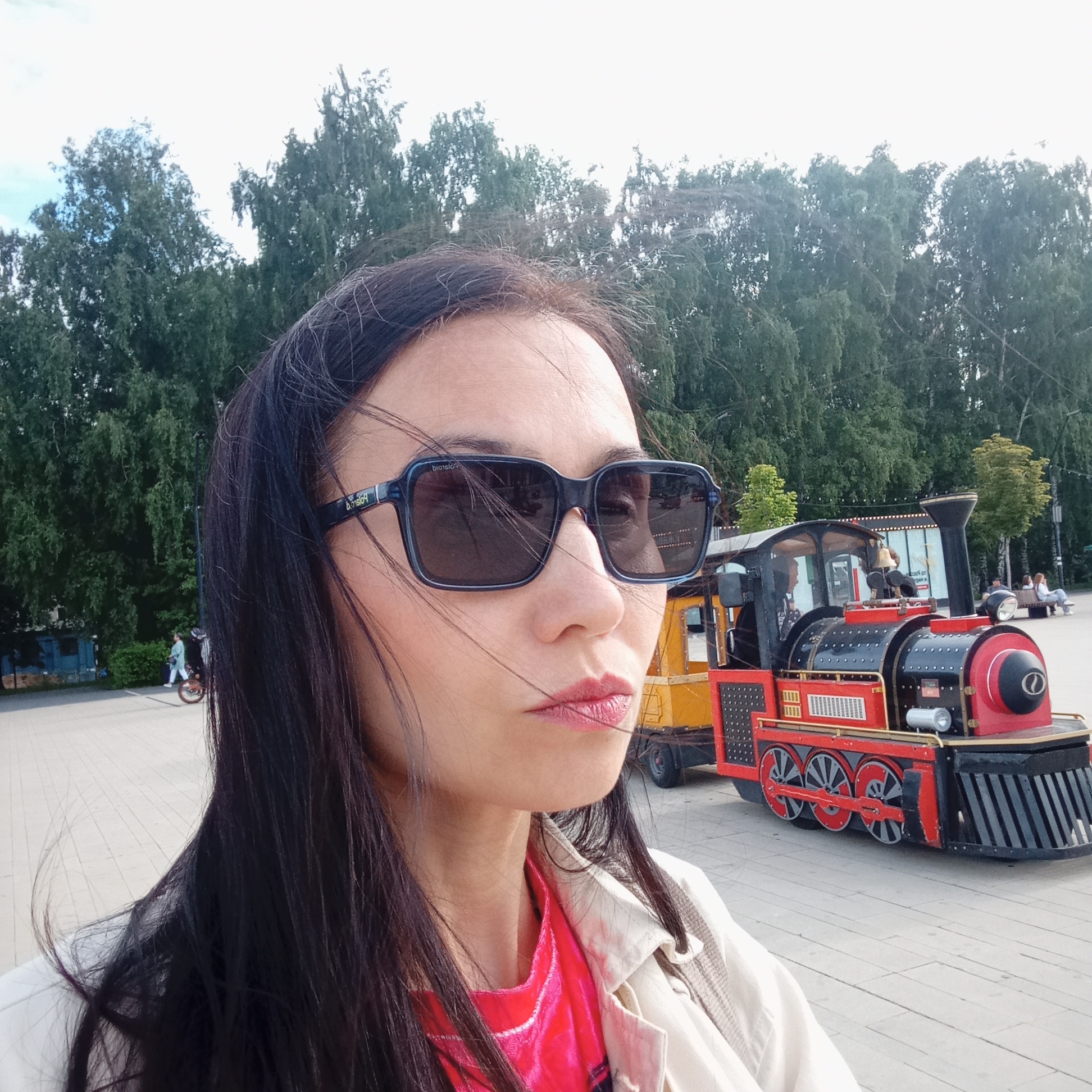 The image captures an adult female in a partial profile, facing right, against a background of a park environment in Izhevsk, Russia. She has long, dark hair, with several strands blowing across her face and forehead, suggesting wind. She is wearing large, dark rectangular sunglasses with "Polaroid" branding visible on the left temple. Her lips are adorned with a vivid pink or magenta lipstick. She is dressed in a light-colored, possibly beige or off-white, jacket or coat over a bright pink or magenta garment.
To her right, prominently featured in the mid-ground, is a red and black miniature train, characteristic of an amusement park ride. The train has a yellow front section, a black smokestack, a bell, red wheels with black spokes, and a cowcatcher-style front. The number "No. 23" is faintly visible on its side. Behind the train, a small dark kiosk or structure with lights is visible, and further back, two individuals are seated on a wooden bench. The ground consists of a wide, light-colored paved area, likely concrete or large pavers, indicating a promenade or park pathway.
In the left mid-ground, a person riding a unicycle is seen in motion on the paved surface, wearing light-colored clothing. Beyond the paved area and behind the main subject, a densely treed area with mature deciduous trees forms the background, topped by a bright, overcast sky. A small blue building or structure is partially visible behind the trees on the far left. The scene depicts a recreational area consistent with a public park or embankment. Given the context of Izhevsk, Russia, the setting strongly suggests the Izhevsk Pond Embankment (Набережная Ижевского пруда), a known recreational zone featuring such attractions and open spaces.
