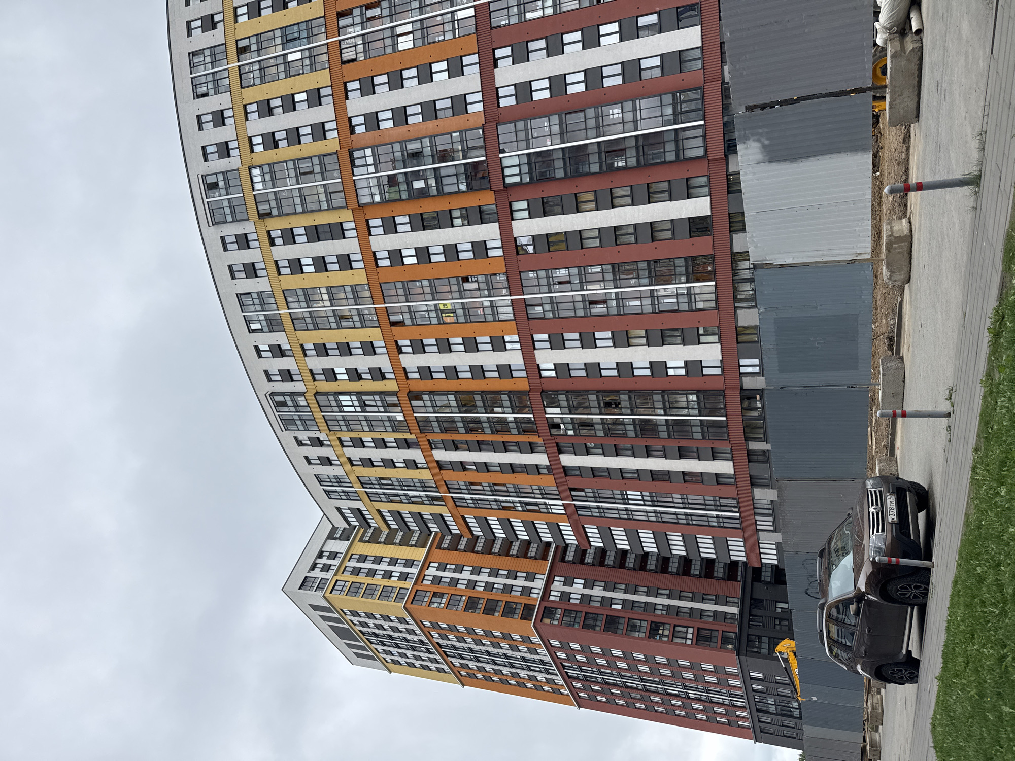 The image captures a modern apartment building in Saint Petersburg, Russia, under an overcast sky. The building is characterized by its colorful facade, with horizontal bands of yellow, orange, white, and brown/red sections, each punctuated by numerous rectangular windows.
To the right of the building is a temporary construction fence made of gray metal panels. A dark-colored SUV is parked adjacent to the fence, with a visible license plate. A utility pole is visible next to the car and fence. A strip of green grass borders the sidewalk on the far right. An arm of construction machinery is visible behind the fence.