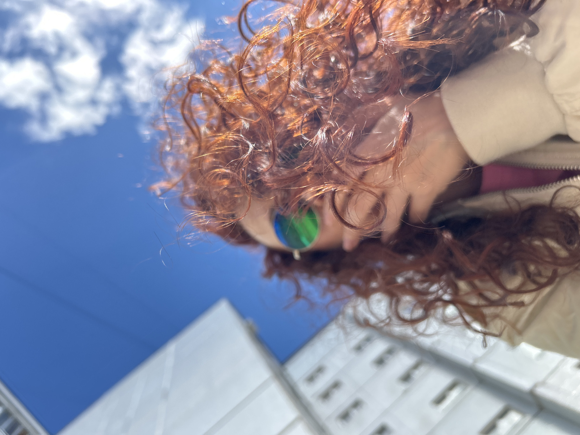 A low-angle shot looks up towards a person and the sky. The person has red, curly hair and is wearing sunglasses with green-blue lenses. They are also wearing a light-colored jacket. Behind the person are buildings and a bright blue sky with some clouds. The buildings are light-colored with a modern architectural style. Based on the information given, this photo was taken in the city of Chaikovsky, Russia.