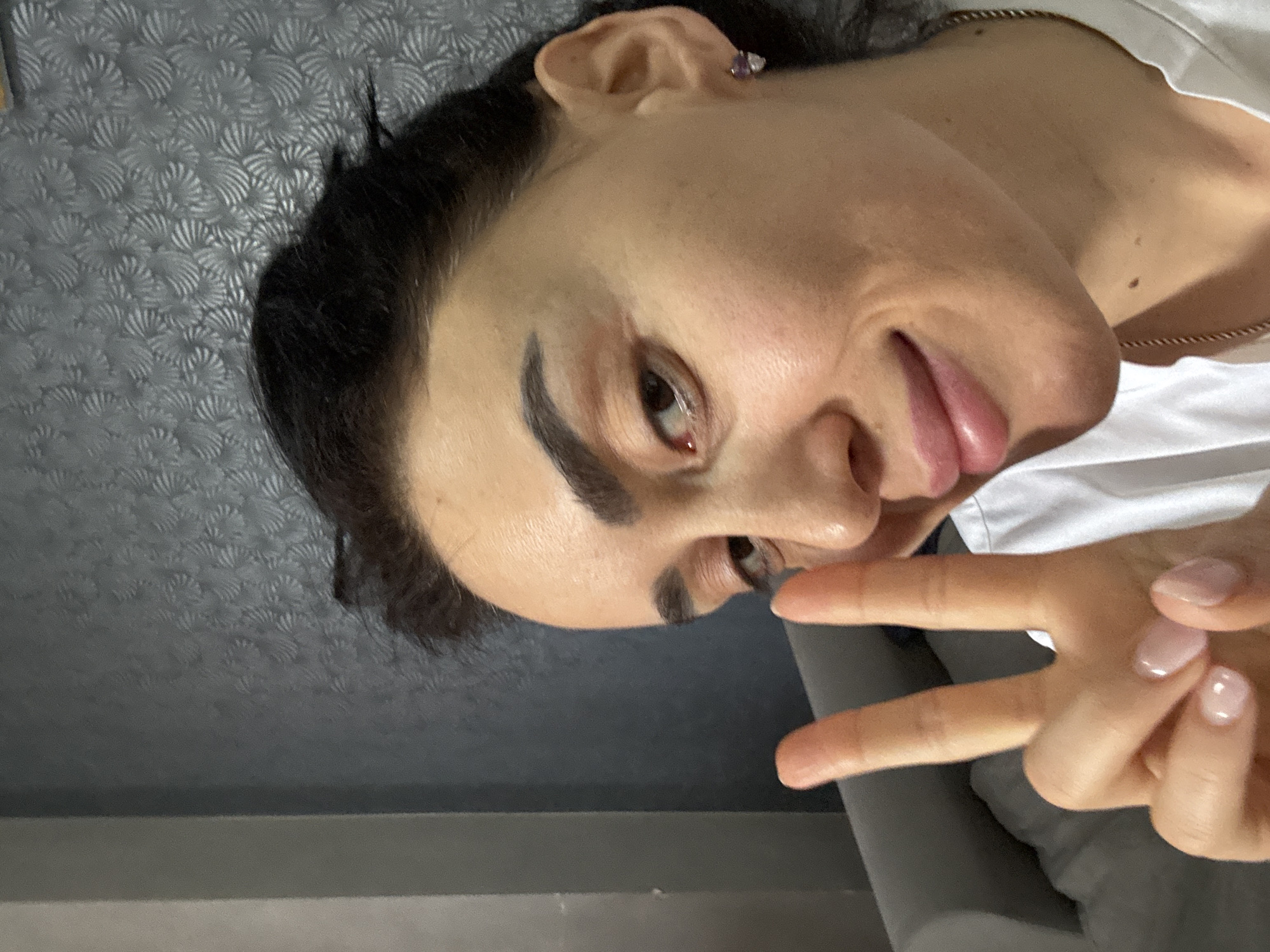 A person with dark hair is shown from a slightly skewed angle, making a peace sign with their hand. The person has fair skin, dark eyebrows, and is wearing a white top. A silver necklace is visible around their neck. The background consists of a gray surface with a subtle textured pattern. Another gray surface is visible below. No specific details indicating the location of Tbilisi, Georgia, are discernible from the image.