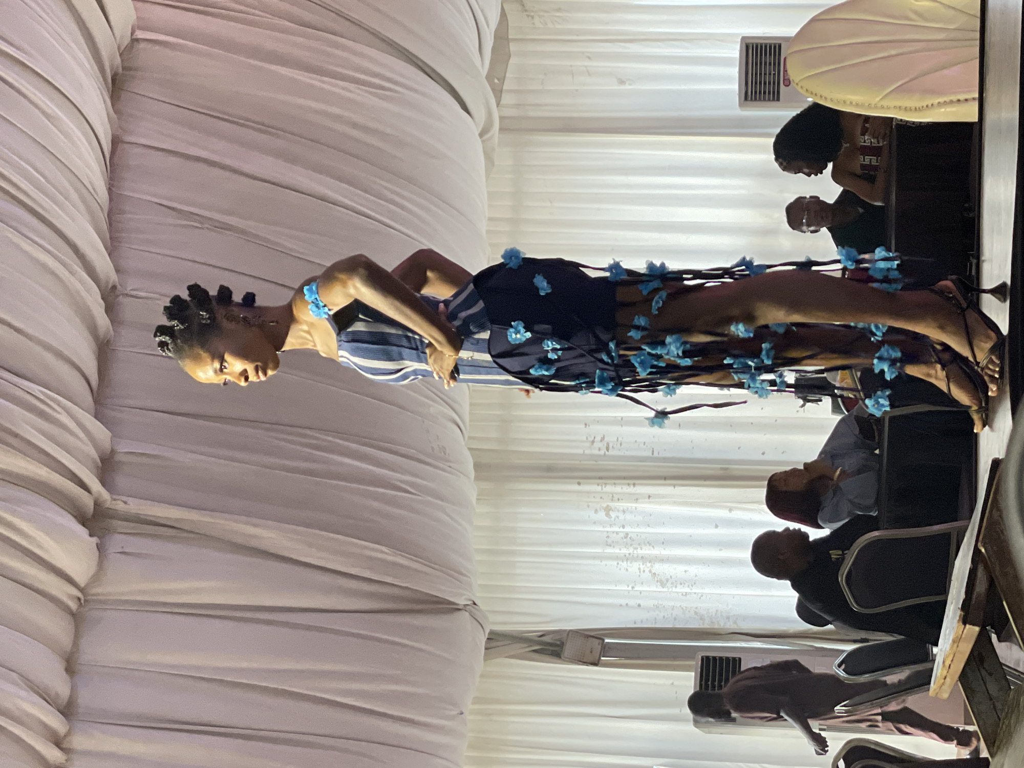 The media file shows a fashion show taking place in Barakin Lerawi, Nigeria. A model is walking down a runway, showcasing a dark blue dress adorned with light blue flowers. She has her hair styled in braids and is wearing blue jewelry. In the background, audience members are seated, watching the show. The setting appears to be indoors, with white drapery covering the walls. A white armchair is visible on the right side of the image. The lighting suggests it is nighttime or the event is taking place in an indoor venue with artificial lighting. The overall mood seems to be one of anticipation and enjoyment, typical of a fashion event. No visible text is present in the media file.