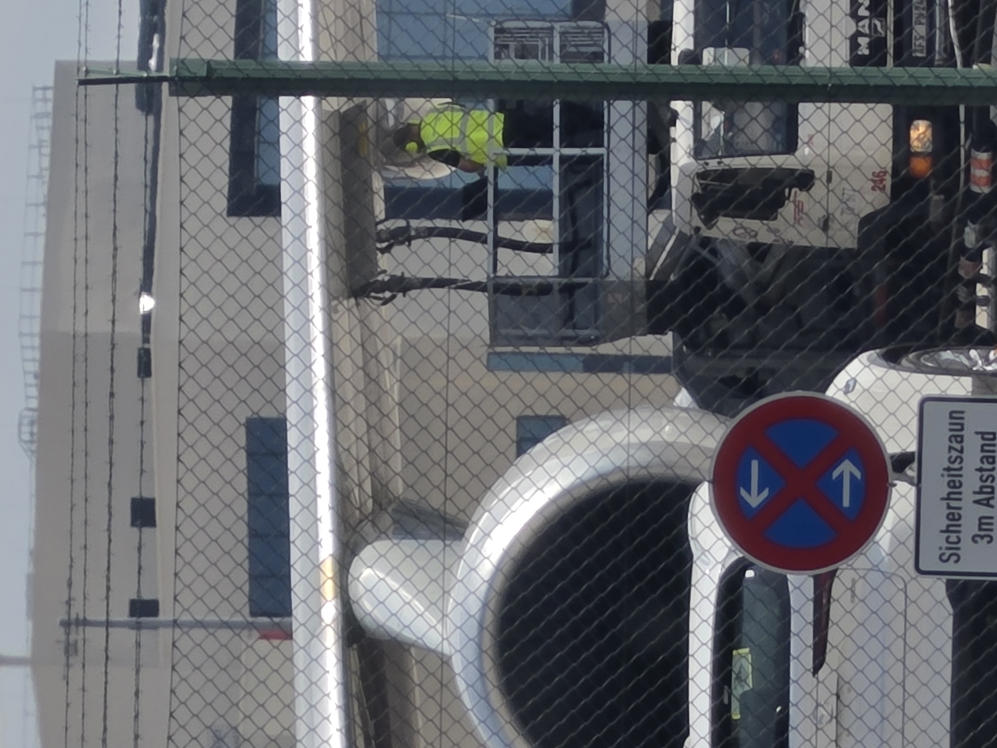 An aerial refueling operation is underway at Frankfurt Airport, Germany. A tanker aircraft's fueling boom is extended towards a refueling station. A worker in a high-visibility vest is operating the refueling equipment from a raised platform. In the foreground, a white car is parked near a "No Parking" sign with German text indicating a 3-meter safety distance. A large truck with the MAN logo is also visible in the background, suggesting it is part of the airport's ground support infrastructure. The scene is viewed through a chain-link fence with barbed wire on top.