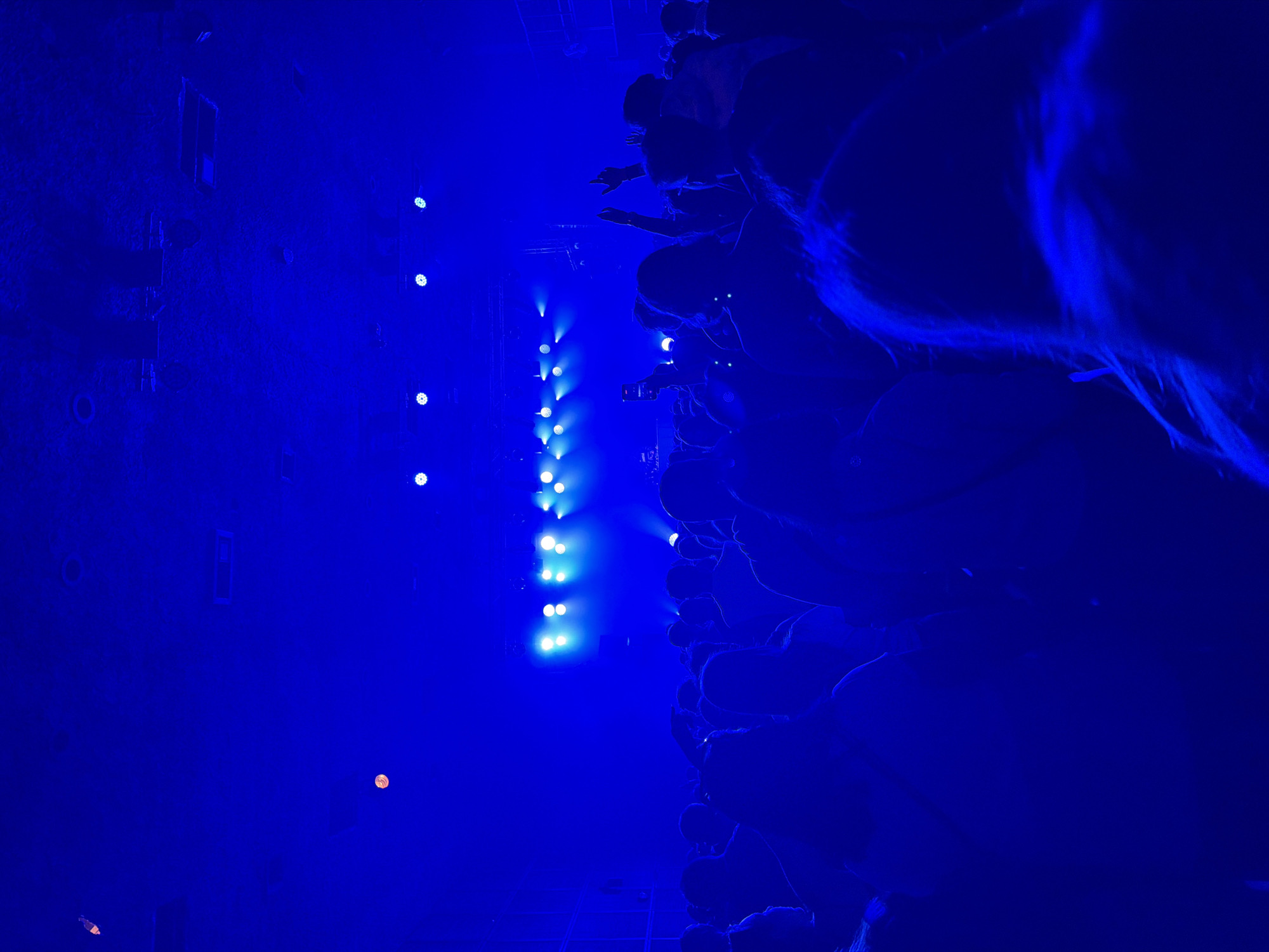 The image depicts a concert setting. A crowd of people faces a stage illuminated by a vertical row of bright lights. The scene is dominated by blue lighting. Details are obscured due to the low light conditions and perspective. The location and city are unknown.