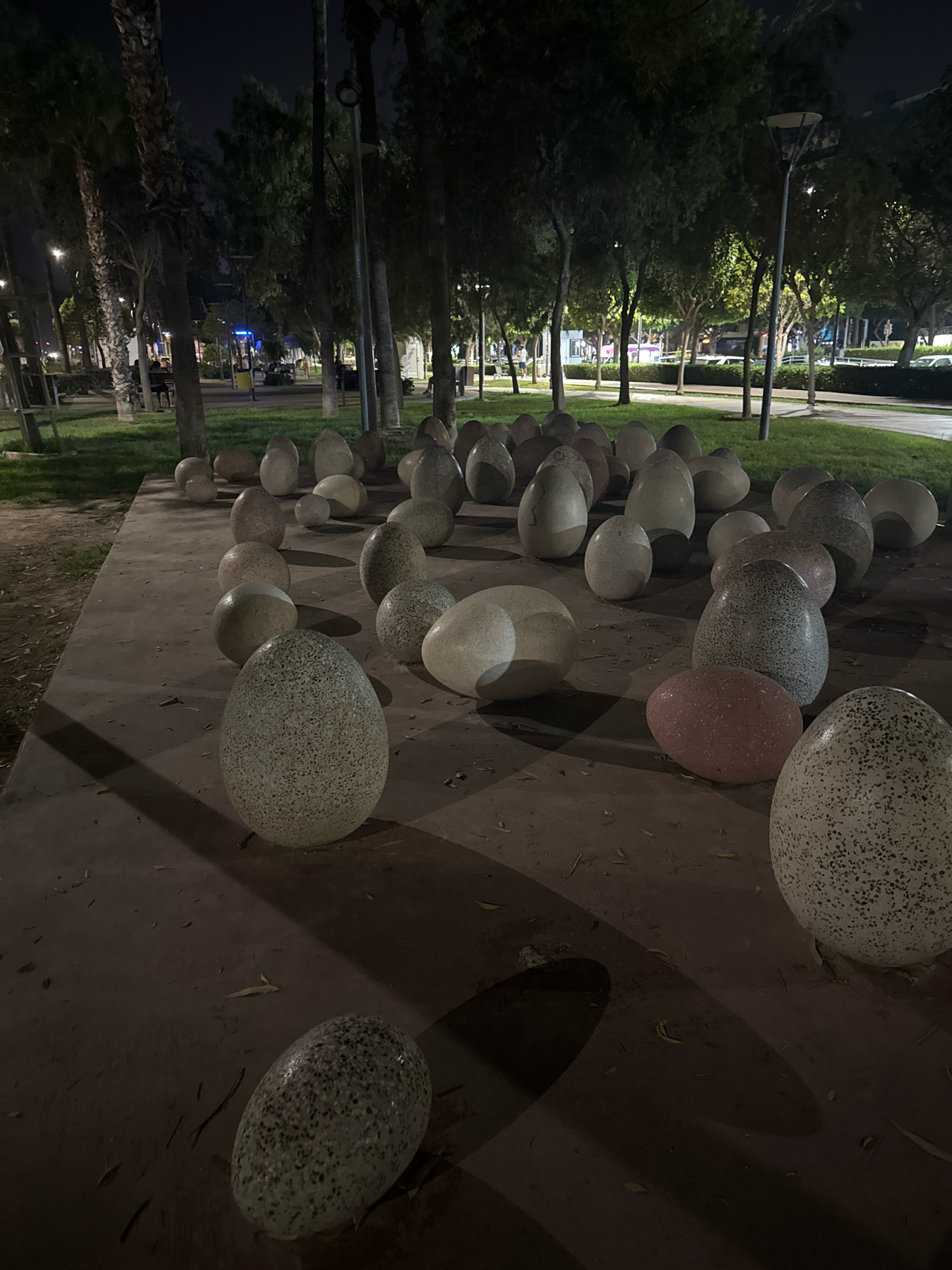 An outdoor nighttime scene in Limassol, Cyprus, features a collection of large, egg-shaped sculptures scattered across a paved area. These sculptures vary in size and color, with some appearing speckled grey and white, while others are a solid pinkish hue. They are arranged in clusters and individually, casting distinct shadows on the ground due to overhead lighting.
In the background, a park setting is visible with mature trees lining a grassy area and a pathway. Streetlights illuminate the scene, creating a somewhat subdued atmosphere. Beyond the park, elements of the urban environment can be discerned, including buildings and what appears to be a road with vehicle headlights. A few individuals are present in the background, seemingly engaged in casual activities, contributing to the overall impression of a public space in use during the evening. The ground is covered with some fallen leaves.