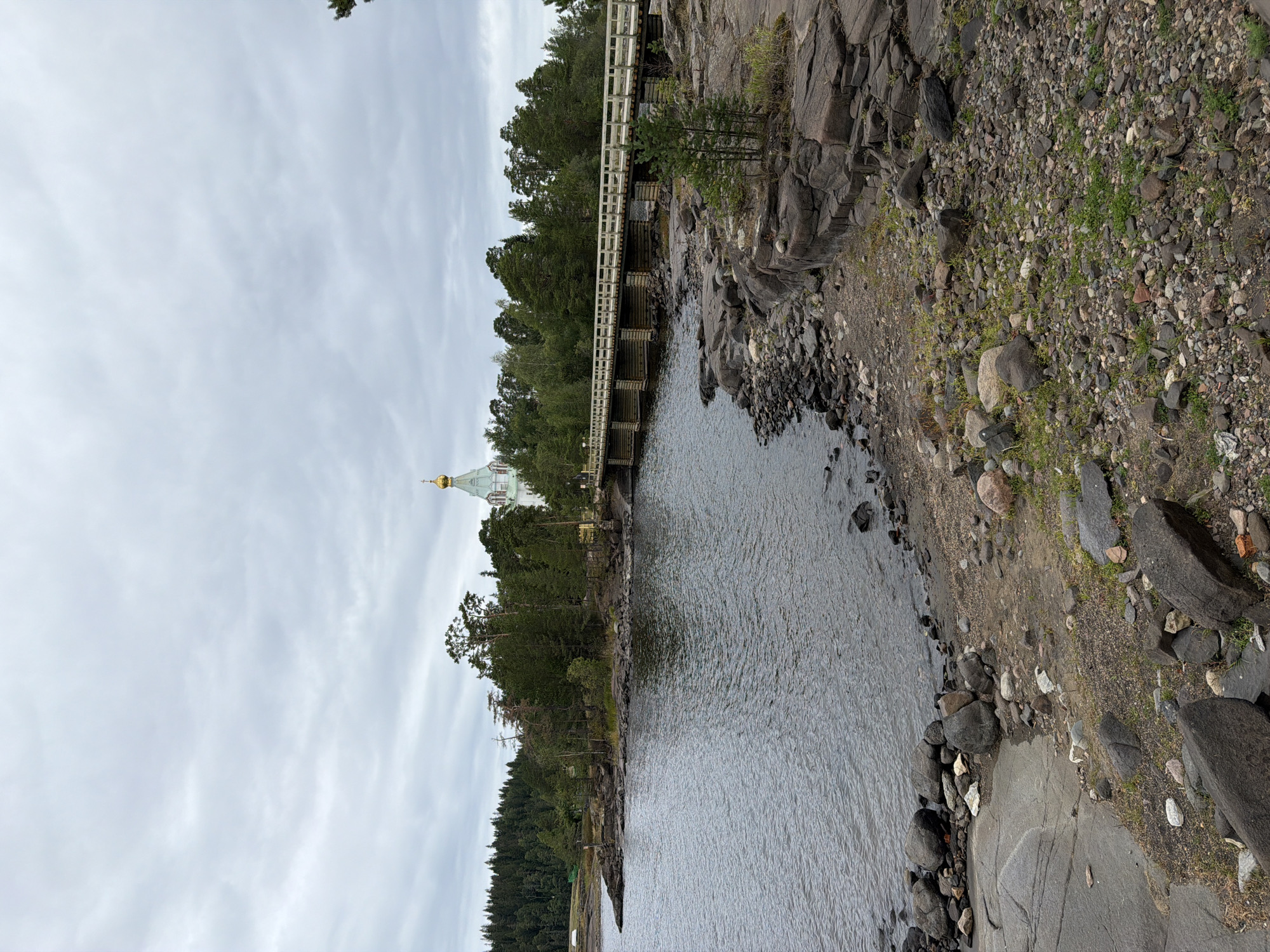 A wooden bridge crosses a dark body of water, connecting two small islands covered in pine trees. On the far island, a white building with a golden dome, likely a church, is visible amongst the trees. The foreground of the image features a rocky shoreline with scattered stones and sparse vegetation. The sky is overcast with gray clouds. The scene appears to be a natural landscape with man-made structures integrated into it.