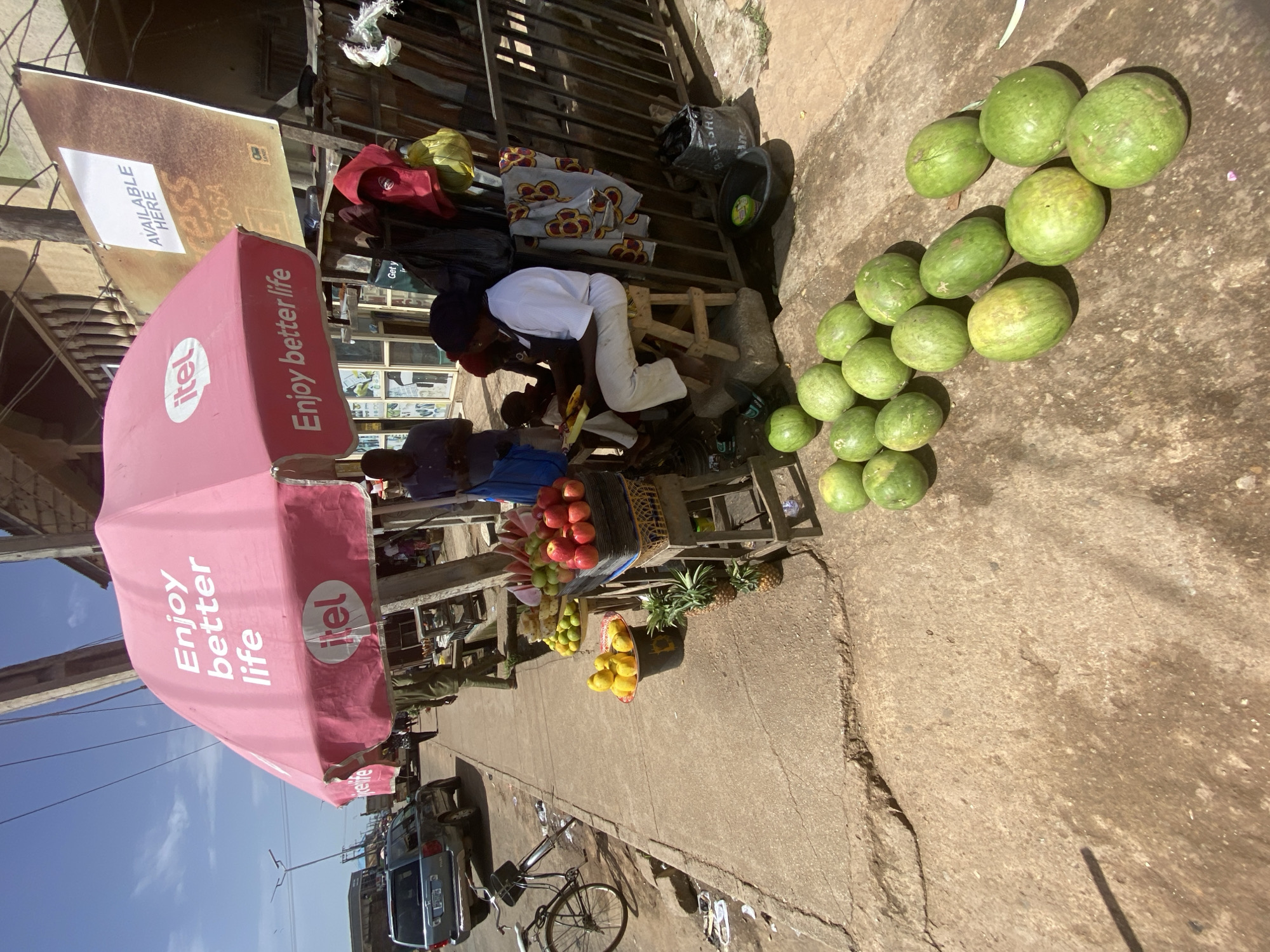 This media file captures a vibrant street scene in Bukuru, Nigeria, likely during the daytime given the clear skies and bright sunlight casting shadows. The foreground features a bustling market stall, adorned with a prominent pink umbrella displaying the "itel Enjoy better life" slogan and logo. Several people are visible, including a vendor or customer in a white shirt and light-colored pants, interacting with goods at the stall.
The stall itself is laden with a variety of fresh produce. Piles of green watermelons are arranged on the ground to the right, while baskets and trays overflow with apples, pineapples, and yellow fruits, possibly mangoes or citrus. Behind the stall, a covered structure with wooden slats suggests a more permanent shop or shelter, with various items, including textiles and bags, visible.
Further in the background, the street continues, with a parked car and a bicycle suggesting the presence of ongoing daily life and commerce. The ground is paved concrete, typical of many urban and semi-urban areas in Nigeria. The overall impression is one of lively commerce and everyday activity within a community setting. The visible text "AVAILABLE HERE" on a sign above the stall provides a practical detail about the services or products offered.