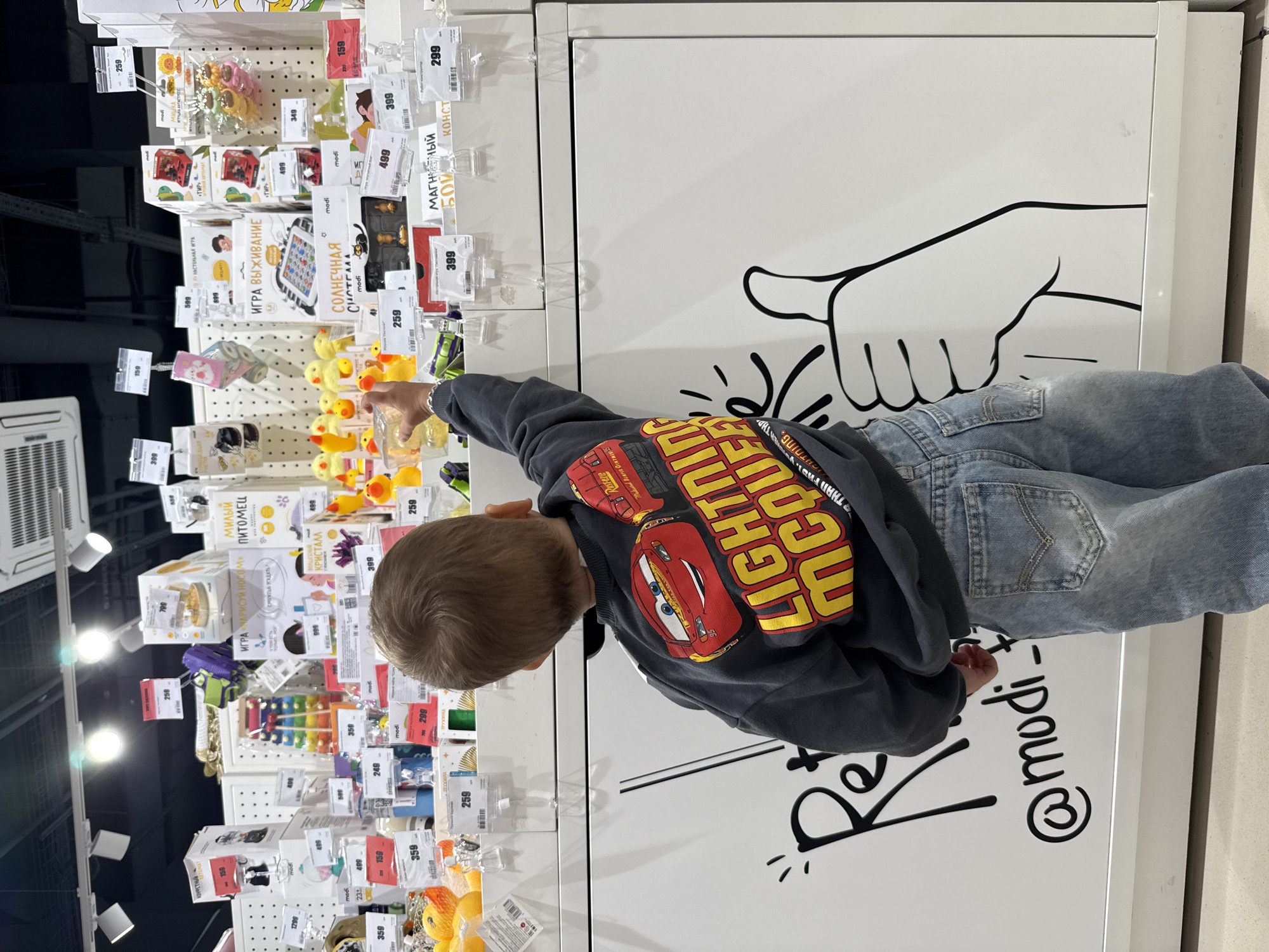 A young boy, viewed from behind, wearing a dark grey sweatshirt with a large graphic of Lightning McQueen from the Disney Pixar movie Cars and light blue denim jeans, stands in front of a brightly lit retail display of toys and games. He is reaching his right hand out towards the merchandise. The display consists of white shelving units with numerous boxes and products, many of which have price tags. Various toys are visible, including what appear to be bath toys, board games, and plush toys. The background shows a store ceiling with recessed lighting. A large graphic of a stylized thumb up and the text "@modi_" are visible on the white surface below the display. The setting is a retail store, possibly in Rusanovo, Russia, given the text on the merchandise and the context provided by the prompt.