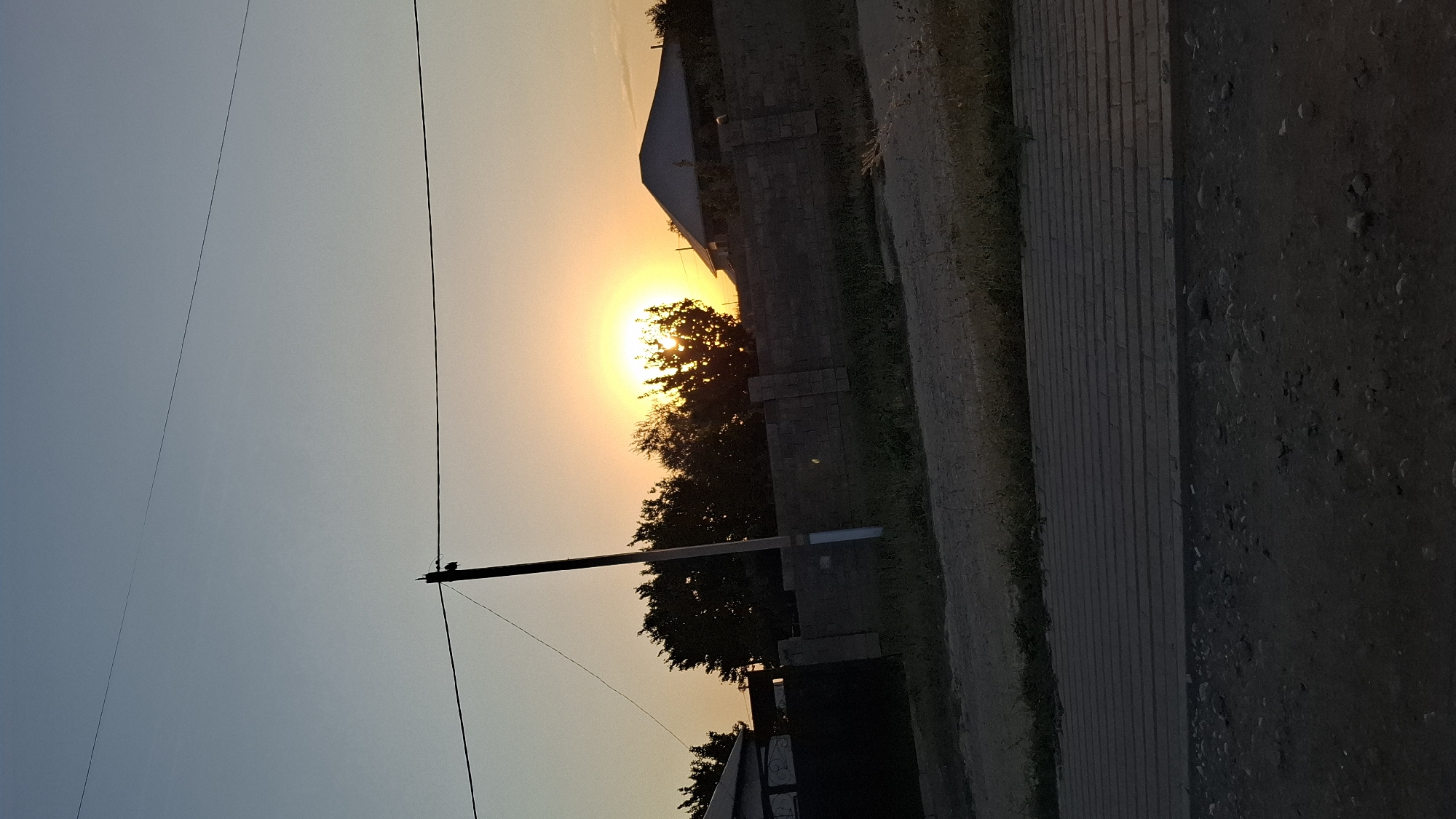The image depicts a sunset scene in the city of Shubarssu, Kazakhstan. The sun is setting behind a line of trees and a brick fence, casting a bright yellow and orange glow across the sky. A silhouette of a utility pole stands to the left of the frame, with power lines extending horizontally across the upper portion of the image. In the foreground, a paved walkway or road is visible, with a dirt or gravel area to its left. Two houses and a black gate are partially visible in the background, behind the fence. The overall impression is one of a rural or suburban residential area at dusk.