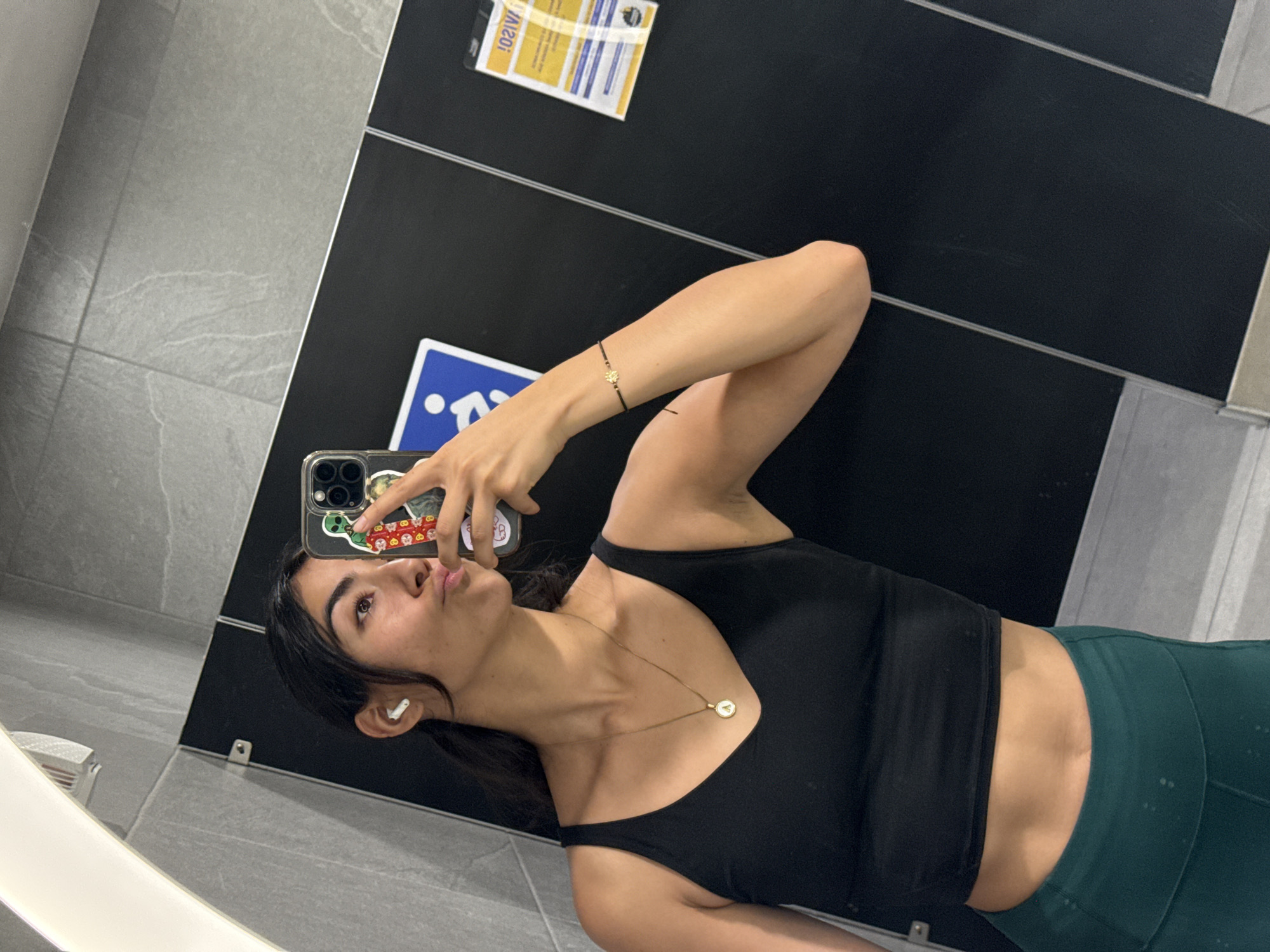A young woman is taking a selfie in a bathroom, presumably at a gym, evidenced by her activewear: a black tank top and dark green leggings. She is holding her phone in her right hand, which has a delicate bracelet on her wrist. Her phone case is transparent with stickers. She is wearing an AirPod in her left ear and a gold necklace. The background consists of a tiled wall with black partitions and informational signs, including one indicating accessibility. The bathroom appears modern with gray tile. There is not enough information to infer the exact location of this bathroom in Puebla, Mexico.