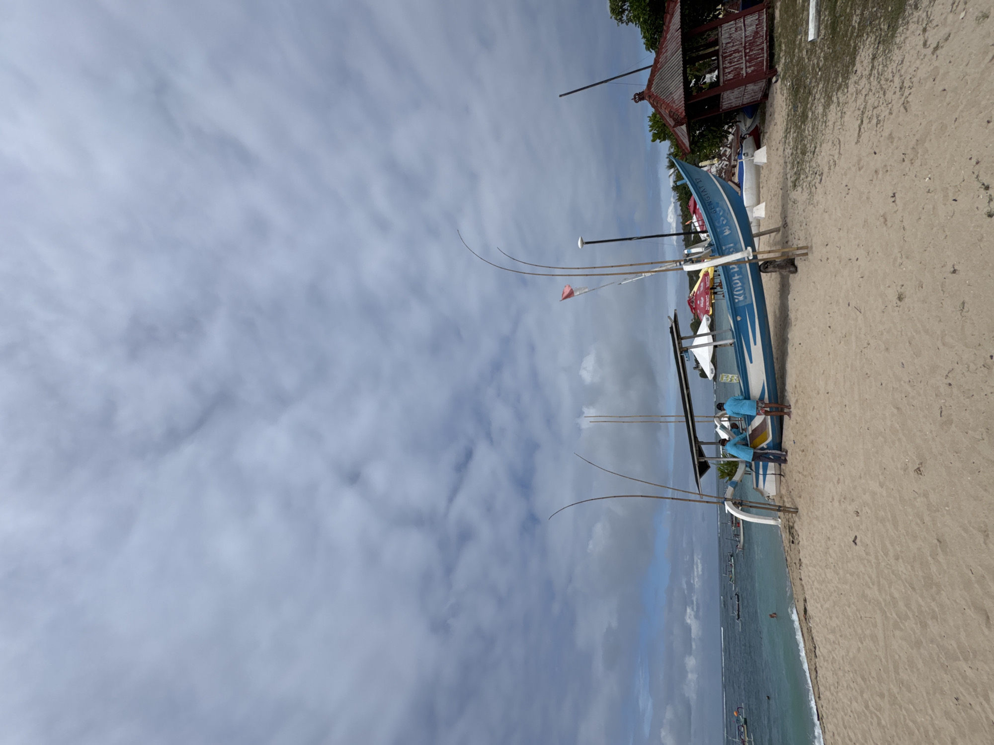 The image depicts a beach scene in Kuta Selatan, Indonesia. A blue and white boat is positioned on the sandy shore, supported by stands. Two individuals are seen standing near the boat. The beach extends towards the ocean, where several boats are visible in the water. The sky is overcast with clouds. A building with a red roof is present near the boats on the sand.