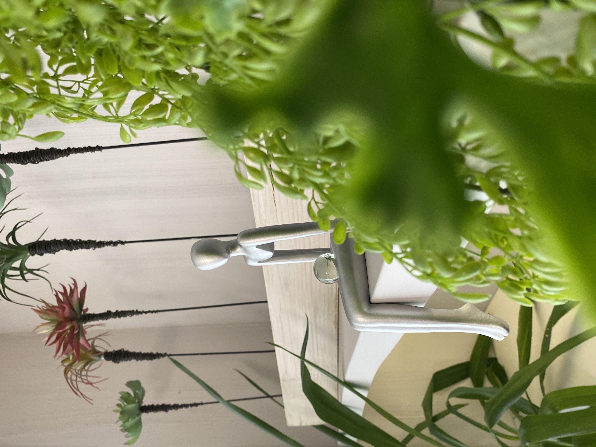 This image depicts an indoor decorative arrangement featuring a minimalist silver-colored figure amidst various artificial plants. The scene is static, showcasing objects rather than an event.
In the center, a stylized, smooth silver figure, resembling a mannequin, is seated on a light wooden shelf. The figure is posed with its knees bent and arms resting on its lap, holding a small, clear spherical object between its hands. The shelf appears to be made of light-colored wood with horizontal grain, and it is positioned against a white paneled wall with distinct horizontal grooves.
To the left of the shelf, three artificial succulents, resembling air plants (Tillandsia), are suspended. Each plant is attached to a thin black rod or wire, which is wrapped in a dark material near the plant base. The plants vary in appearance: the uppermost one is green, the middle one features a striking reddish-pink center with green outer leaves, and the lowest one is entirely green.
The foreground and parts of the background are dominated by blurred, vibrant green foliage, likely artificial vines, creating a sense of depth and framing the central display. This blurred green mass suggests it's closer to the camera, partially obscuring the wider scene. The overall lighting is bright and diffused, typical of an indoor setting during the daytime. There is no visible text in the image. The location is an interior space, possibly a home or office, designed for aesthetic appeal.