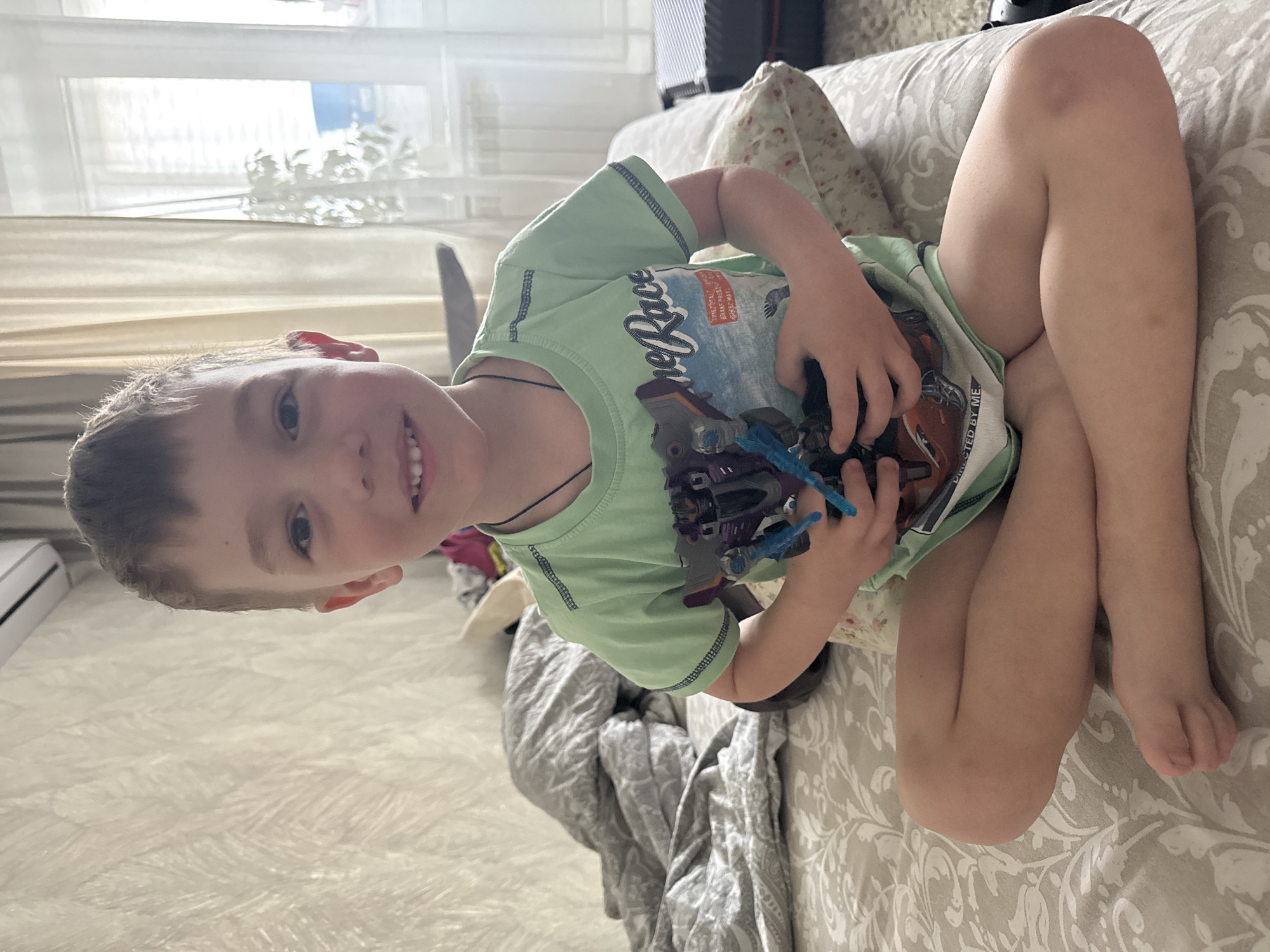 A child is seated on a light-colored patterned sofa. The child, who appears to be a boy, is wearing a green t-shirt with a print and holding a toy. The background includes a window with sheer curtains, and a glimpse of foliage outside. The environment suggests an indoor setting, possibly a home. Based on the prompt, the location is presumed to be in the city of Chaikovsky, Russia.