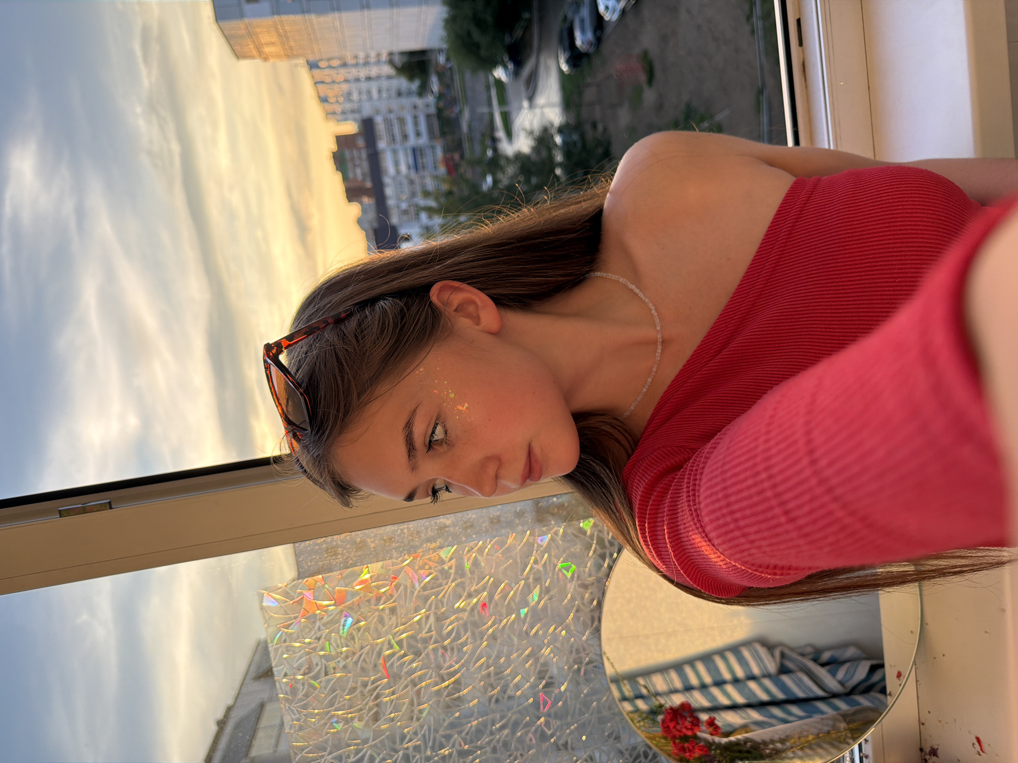 The image shows a young woman with long brown hair, wearing a red off-the-shoulder top and a delicate necklace. She has glitter on her cheek and is wearing sunglasses pushed up on her head. She is looking off to the left, with a thoughtful expression. She is positioned in front of a window that overlooks a cityscape. The window has decorative film on it, which refracts light. In the background, there are apartment buildings and trees, with a cloudy sky above. The scene appears to be set during either sunrise or sunset, given the warm light. The overall impression is one of a casual, personal moment captured against an urban backdrop. The location is identified as Chaikovsky, Russia.