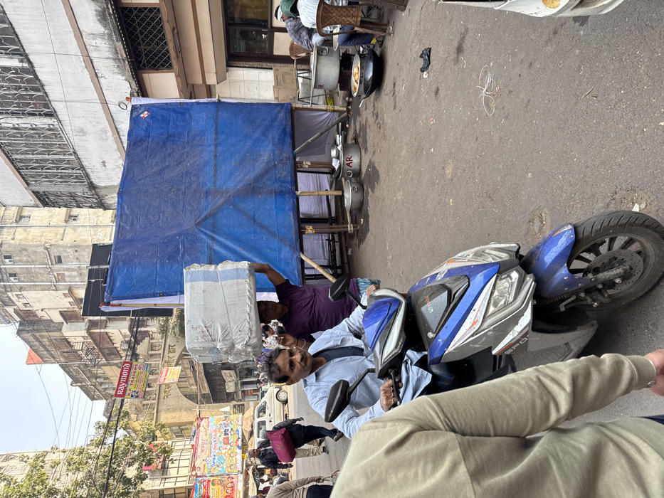 A blue scooter is on the street, being driven by a man in a light blue shirt. Another man is behind him, holding up a large, wrapped package. Behind them is a food stall with a blue tarp overhead and large cooking pots on a makeshift table. Buildings and street signs are visible in the background. The scene is shot from the perspective of a person standing close by, their arm and jacket partly in the frame.