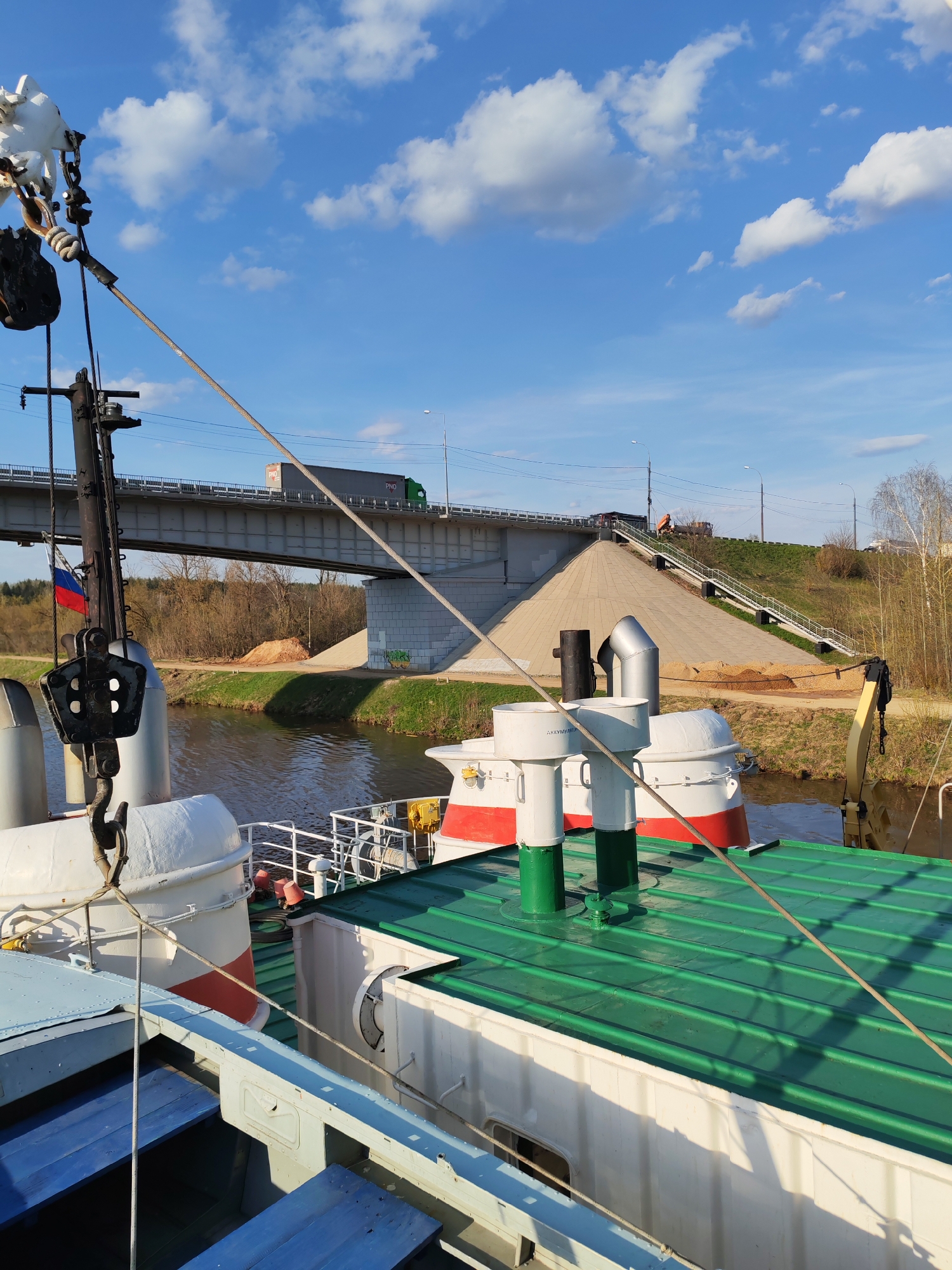 The image depicts a view from the deck of a river vessel navigating a canal near Podosinki, Russia. A concrete bridge spans the canal, with a green and gray semi-trailer truck driving across. The canal banks are earthen with sparse vegetation. The river vessel has a green roof, white structures with red striping, and black exhaust pipes. Rigging and tackle are visible on the forward part of the boat. The sky is blue with scattered white clouds.
