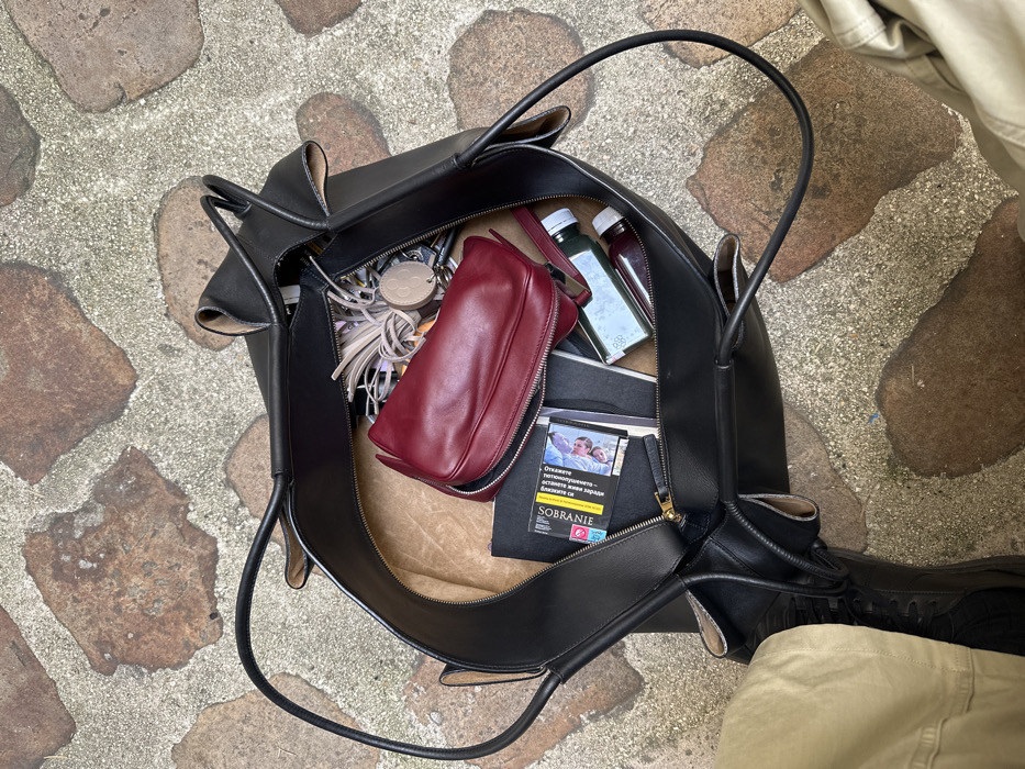 An elevated view depicts an open black leather tote bag resting on irregular reddish-brown stone pavement with grey mortar. The scene is located in Paris, France, identifiable by the French-language health warning on a Sobranie cigarette pack within the bag. The bag's interior contains a dark red zippered pouch, the aforementioned Sobranie cigarette pack (featuring an image of a couple), a black notebook, two small bottled beverages (one green, one reddish-purple, with "PGD" visible on the green bottle's label), and a cluster of keys attached to multiple keychains, including a light-colored fringed tassel and a round beige disc. Partial views of a person's legs, clad in khaki trousers and dark shoes, are visible at the bottom left and right of the frame, indicating the person is standing over the bag.