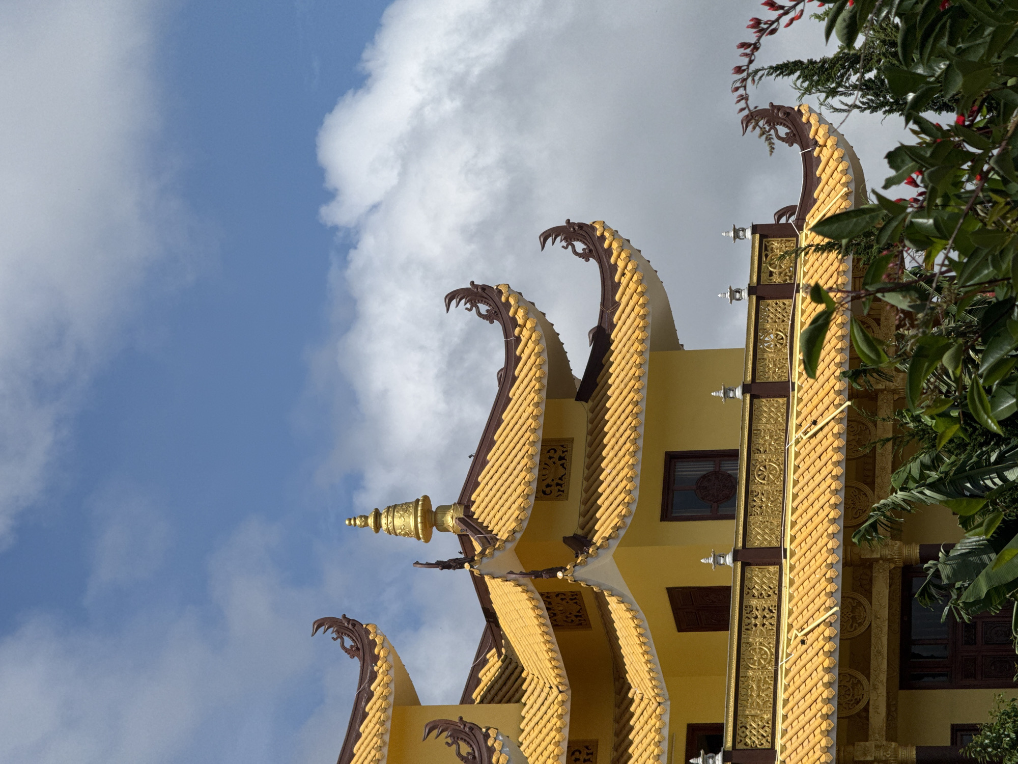 The image shows a close-up view of the yellow, multi-tiered roof of a Buddhist temple, possibly the Truc Lam Zen Monastery, located in Da Lat, Vietnam. The intricate, curved roof tiles are a vibrant yellow, with dark brown trim and decorative elements resembling dragon heads adorning the eaves. A golden spire tops the highest visible section of the roof. The building features ornate golden railings with Buddhist symbols, and dark wooden windows. Lush green foliage, including broad leaves and red-flowered branches, frames the bottom and right sides of the image, obscuring the lower parts of the structure. The sky above is a bright blue, dotted with fluffy white clouds. The scene is well-lit, suggesting it was taken during the daytime. There are no people visible in the image.