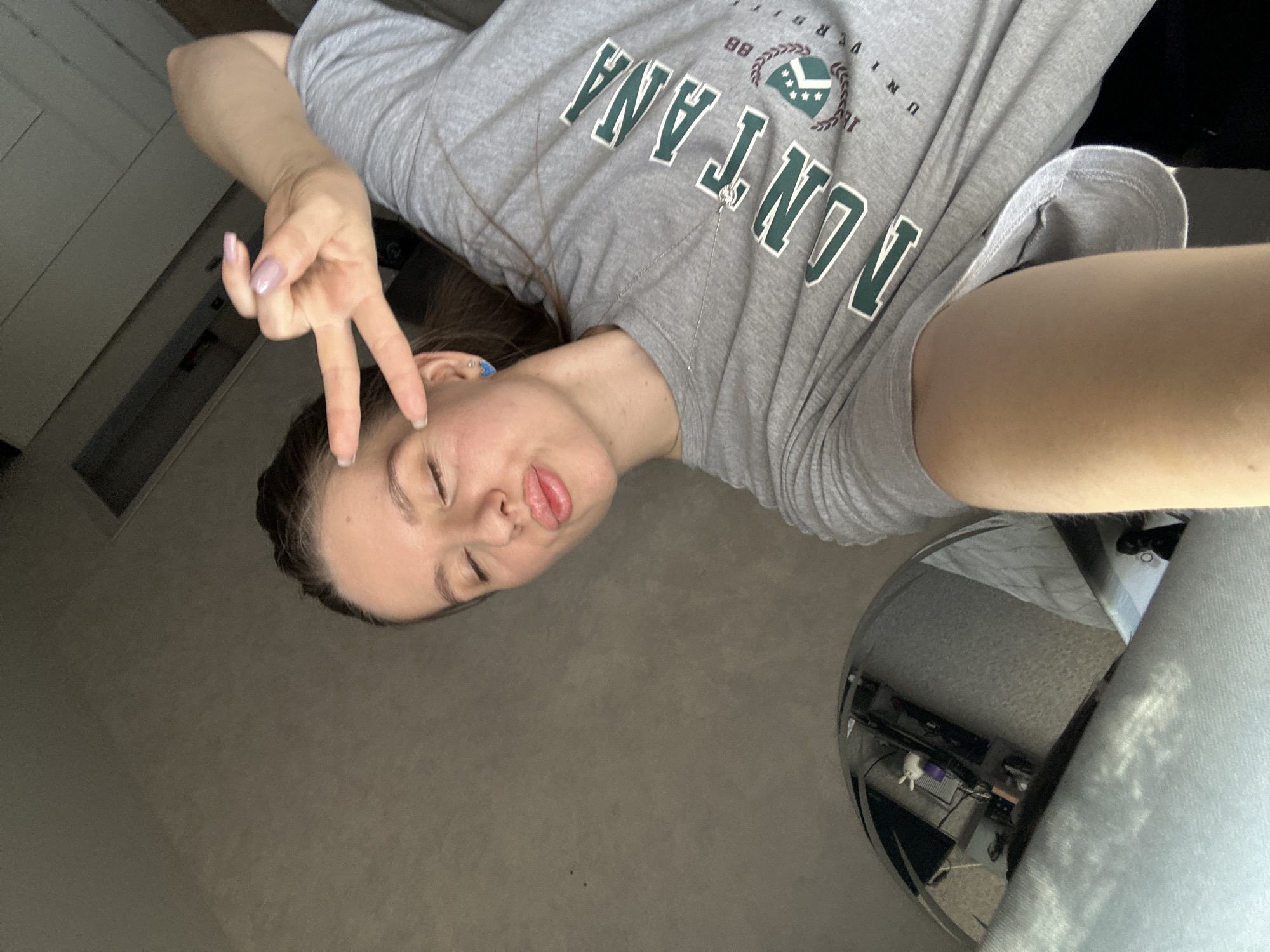 A young woman poses for a selfie. She wears a gray t-shirt with "MONTANA UNIVERSITY 88" printed on the front. She is holding up a peace sign with her left hand and puckering her lips. The background shows a room with a grey carpet and a chrome-colored round table, and there is a reflection in the table of some other furniture and objects. There is no evidence to suggest the location is in the city of Klin, Russia.