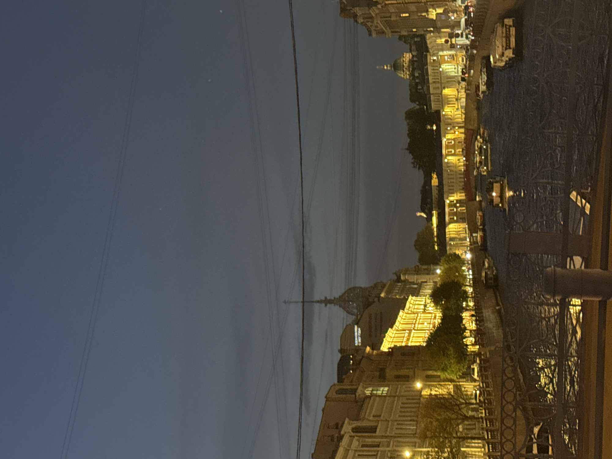 The image depicts a nighttime or late twilight cityscape of Saint Petersburg, Russia, viewed across a body of water, likely a canal or river. A decorative metal railing, possibly part of a bridge or embankment, is visible in the foreground on the right.
The sky, dark blue-grey and appearing overcast, occupies the left portion of the frame and is crossed by multiple overhead wires. On the opposite bank, a series of historic, classical-style buildings are brightly illuminated, casting warm, yellow light that reflects vibrantly on the dark water below. Several boats can be seen on the waterway.
In the distance, prominently silhouetted against the dim sky, is the distinctive spire of the Admiralty building, a key landmark of Saint Petersburg. Other domed structures and architectural details are also visible along the illuminated skyline.
No people are discernible in the scene. Faint Cyrillic text is visible on the facade of one of the illuminated buildings, partially reading "БИБЛИОТЕКА" (Library). The overall atmosphere is serene, showcasing the city's architectural beauty under artificial light.