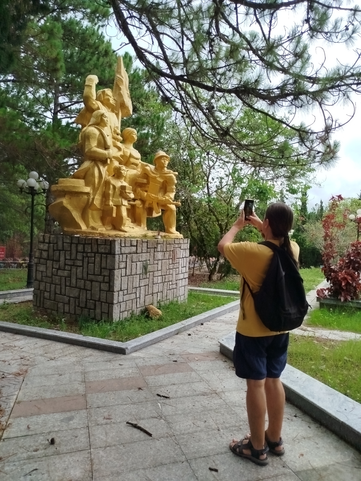 A person is captured from behind, holding a smartphone to photograph a large, golden-colored monument in an outdoor setting. The individual wears a yellow t-shirt, dark blue shorts, and dark sandals, with a black backpack. Their hair is tied back.
The monument features multiple figures, including adults and children, carved in a dynamic pose. One central figure appears to raise an arm while another holds a flag or banner. Other figures seem to hold tools or a rifle, suggesting a commemorative theme of unity, labor, or a historical event. The statue is mounted on a rectangular base constructed from textured grey stone blocks arranged in a basketweave pattern.
The scene is set in a park or memorial area, consistent with the provided location of Cam Ly - Đà Lạt, Vietnam. Tall pine trees with slender needles dominate the background and frame the top of the image, their branches reaching towards an overcast or cloudy sky. Lush green grass surrounds the monument's base, and landscaped bushes, some with striking reddish leaves, are visible in the background. A paved walkway of grey and reddish-brown tiles leads towards the monument. A street lamp with three spherical lights stands to the left of the statue. Pine needles and small twigs are scattered on the ground, indicating a natural, arboreal environment. The lighting suggests it is daytime, likely a cloudy day. A small, obscured red sign with illegible white text is partially visible behind the lamp post.