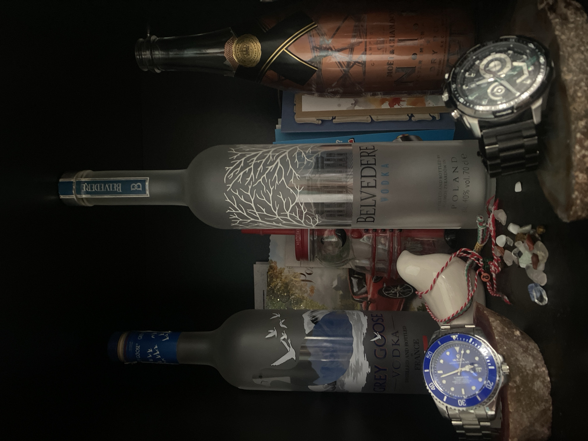 A still life photograph features three liquor bottles, two wristwatches, and a ceramic bird on a dark shelf. On the left is a frosted glass bottle of Grey Goose vodka, adorned with an illustration of flying geese. Next to it is a frosted glass bottle of Belvedere vodka with a white, tree-like design. To the right is a bottle of Moët & Chandon champagne with a copper-colored label and black ribbon detailing.
A blue-faced wristwatch with a silver band is positioned on the left, resting on a piece of rough, brown material. A second wristwatch with a black band and a complex, multi-dial face is on the right, also on similar brown material.
A small, white ceramic bird is placed between the vodka bottles. It is adorned with a traditional Bulgarian martenitsa, a red and white string with beads and a small black "8" charm. Scattered around the bird are small, translucent stones. In the background, partially obscured by the bottles, are several books or cards. A red sports car is visible on one of the cards. The overall lighting is dim, with a spotlight effect on the foreground objects.
The presence of the martenitsa suggests a possible connection to Bulgarian traditions, and if this image were to be associated with Plovdiv, Bulgaria, it could be a personal collection displayed in a home or a private establishment within the city.
