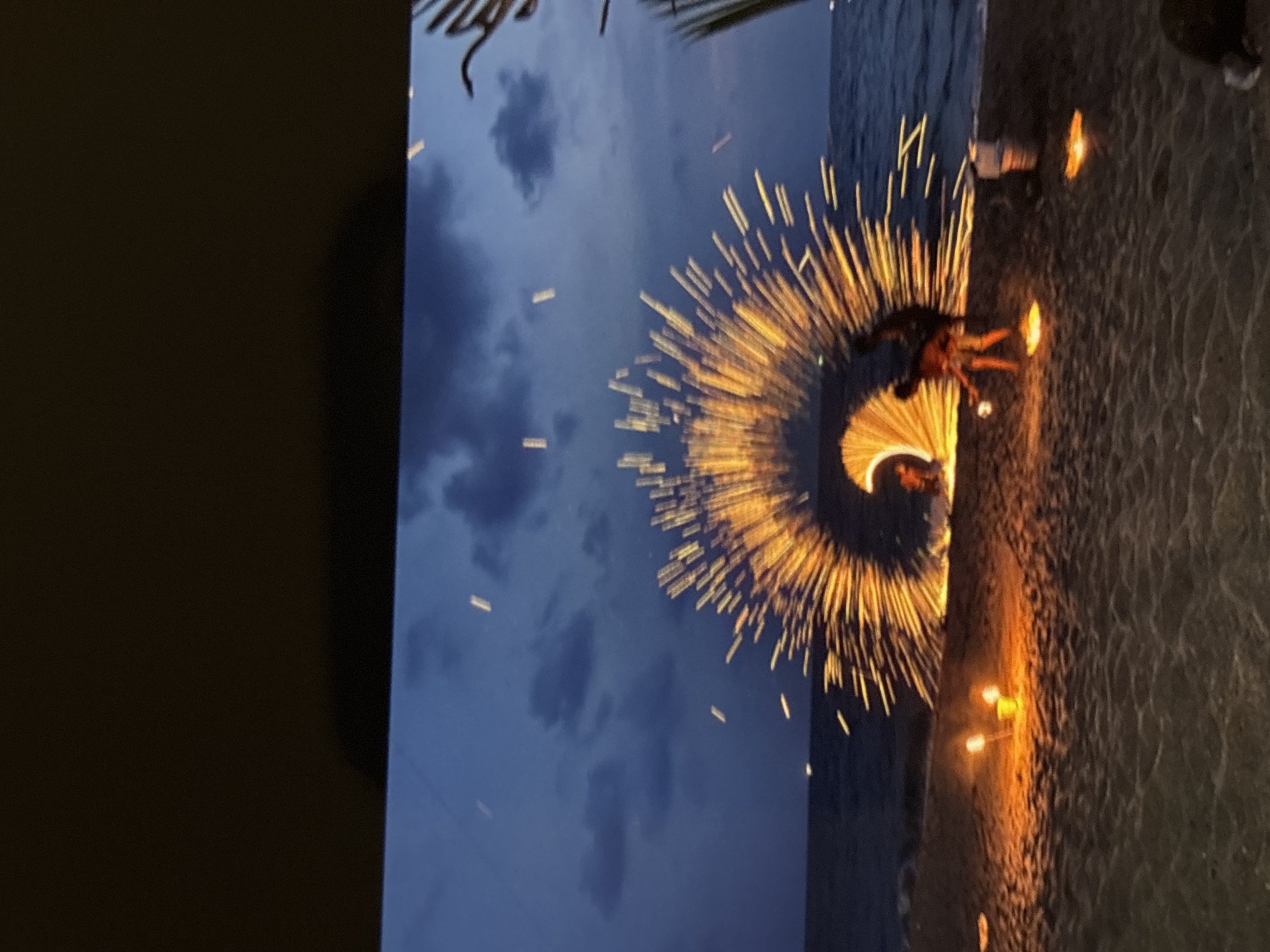 A fire dancer performs on the beach at dusk in Chalong, Thailand. The performer is spinning a fiery object that creates a halo of sparks in the air. The ocean is visible in the background, with waves gently lapping the shore. Several smaller fires are also lit on the sand, casting a warm glow. The sky is a deep blue, with a few scattered clouds. The scene is dimly lit, with the fire being the primary source of light.