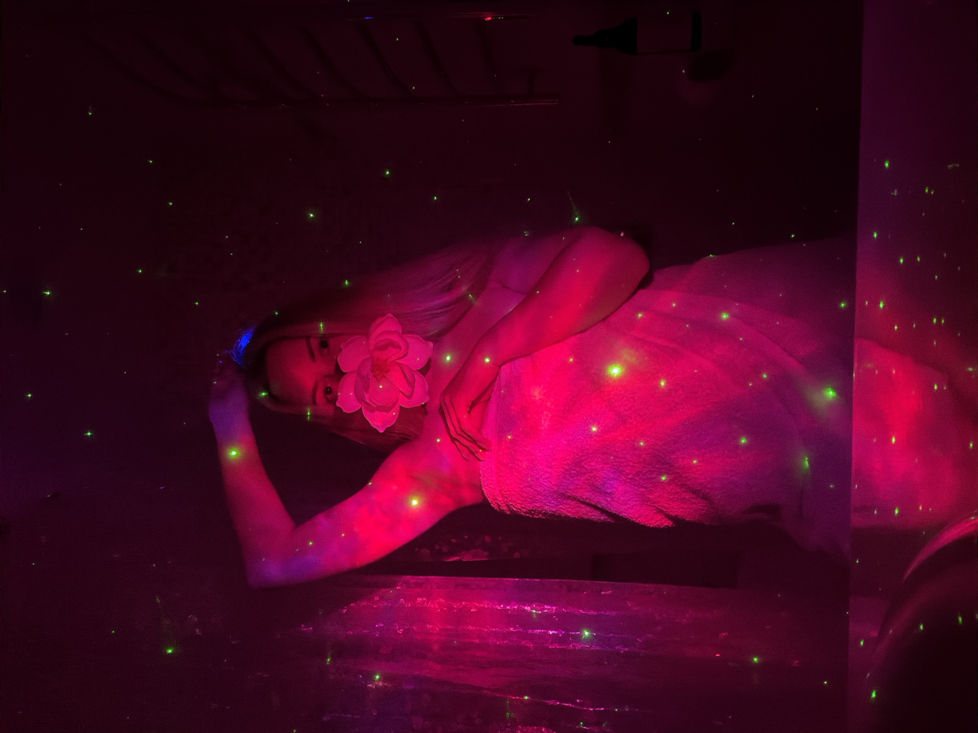 This image captures a person, likely female, with blonde hair, partially visible in a dramatically lit indoor setting. The scene is dominated by vibrant pink and purple ambient lighting, which casts a strong glow on the subject and the immediate surroundings. Numerous small, bright green dots, resembling laser projections or illuminated particles, are scattered across the entire frame, creating a dynamic, almost starry effect.
The individual is positioned on their side, with their head slightly tilted. A large, delicate light pink or white flower is held up to their face, partially obscuring their mouth and nose. Their eyes are visible, looking towards the viewer. The person appears to be wrapped in a white or light-colored towel, which is intensely illuminated by the pink light, covering their body from the waist downwards. One arm is extended, and the other hand rests near their waist. The surface beneath them, and possibly an adjacent wall, reflects the rich pink light. In the upper background, faint outlines of dark objects, such as a shelf or wall decor, are discernible, along with a dark bottle on a higher surface.
The activity appears to be a posed photoshoot, emphasizing atmosphere and aesthetics. The intense, colored lighting and scattered green points suggest a nighttime or artificially darkened indoor environment, possibly a spa, an artistic studio, or an event space. The overall mood is serene, dreamlike, and enchanting, focusing on beauty and artistic expression. No visible text is present in the image. The location context is Votkinsk, Russia.