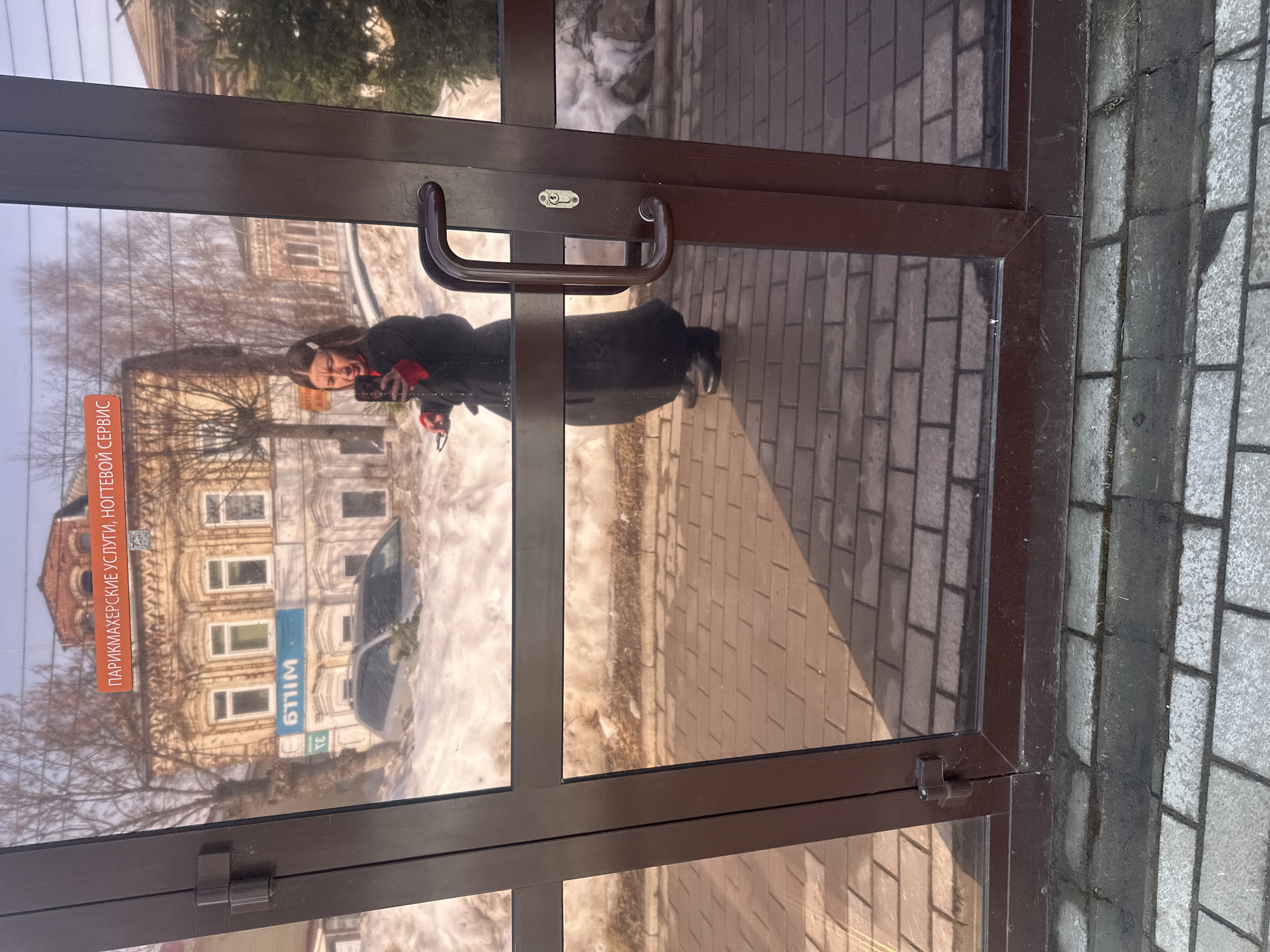 The image captures a reflection in a brown-framed glass double door, revealing an outdoor urban scene in Votkinsk, Russia, during a period of snow accumulation, likely late winter or early spring.
A female individual is prominently reflected, positioned slightly right of center, holding a black smartphone with her right hand, pointed towards herself in the act of taking a photograph or video. She has long dark hair, partially held back by a white barrette, and is dressed in a dark, long coat with a visible red garment underneath. Her left hand holds a pair of eyeglasses.
The reflected background features a light-colored, ornate two-story building with multiple windows and a small rooftop turret, located across a street. An orange banner with Cyrillic text "ПАРИКМАХЕРСКИЕ УСЛУГИ, НОГТЕВОЙ СЕРВИС" (Hairdressing services, nail service) is affixed to the building's facade. A blue sign below the windows displays mirrored Cyrillic text. Significant snow banks line the street, partially obscuring a silver/grey station wagon or SUV parked along the curb. Bare deciduous trees and an evergreen are visible in the background under a clear sky.
The actual foreground shows the dark brown frame of the glass door, which includes a curved handle and a keyhole on the right panel. The ground immediately in front of the door is paved with reddish-brown rectangular bricks, exhibiting clear shadows cast by direct sunlight. Below this brick area, a strip of darker grey paving stones is visible.