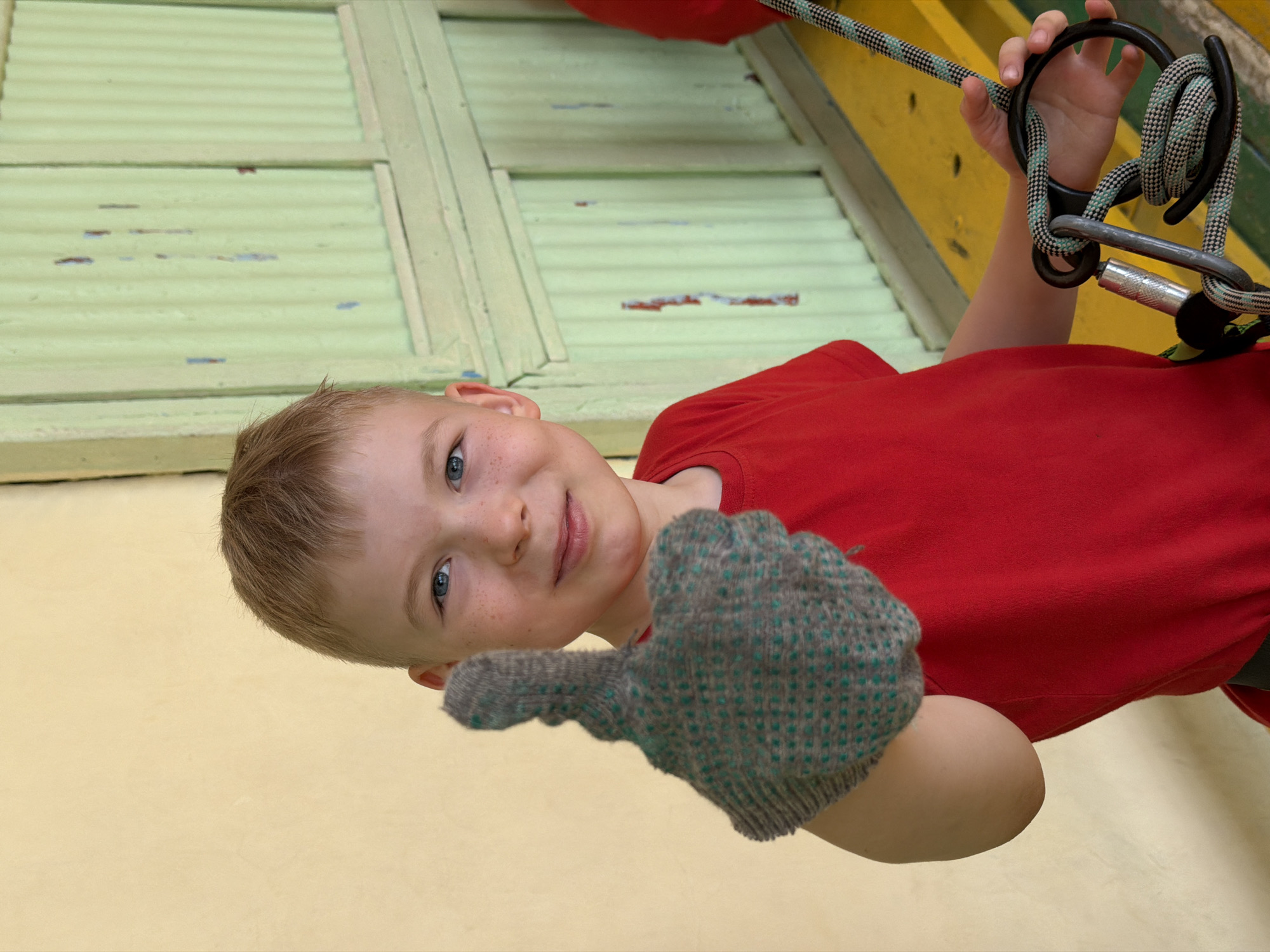 A young boy with blond hair and blue eyes gives a thumbs-up while holding climbing equipment. He wears a red t-shirt and a gray glove with green accents. The climbing equipment consists of a rope, carabiner, and other metal components. The background includes a green and yellow wall, suggesting an indoor climbing facility. There is no information to indicate if this is Izhevsk, Russia.