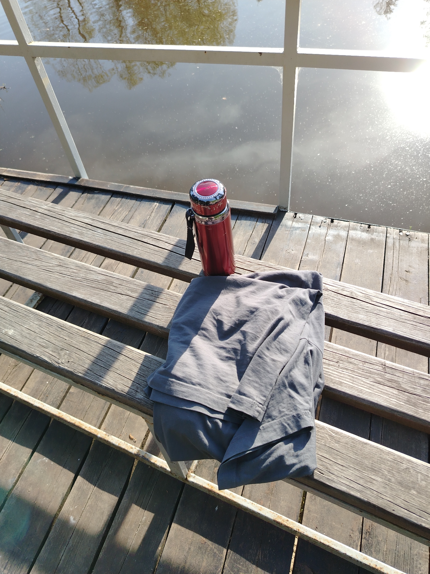 The image shows a scene on a wooden platform or deck next to a body of water in Stroitel', Russia. A red thermos bottle is positioned next to a folded gray shirt on the deck. The platform appears to be part of a walkway or pier, with a white railing partially visible in the background. The water reflects the sky and surrounding trees, with sunlight glinting off the surface. The scene is captured in daylight, with shadows cast by the sunlight across the wooden planks of the platform.