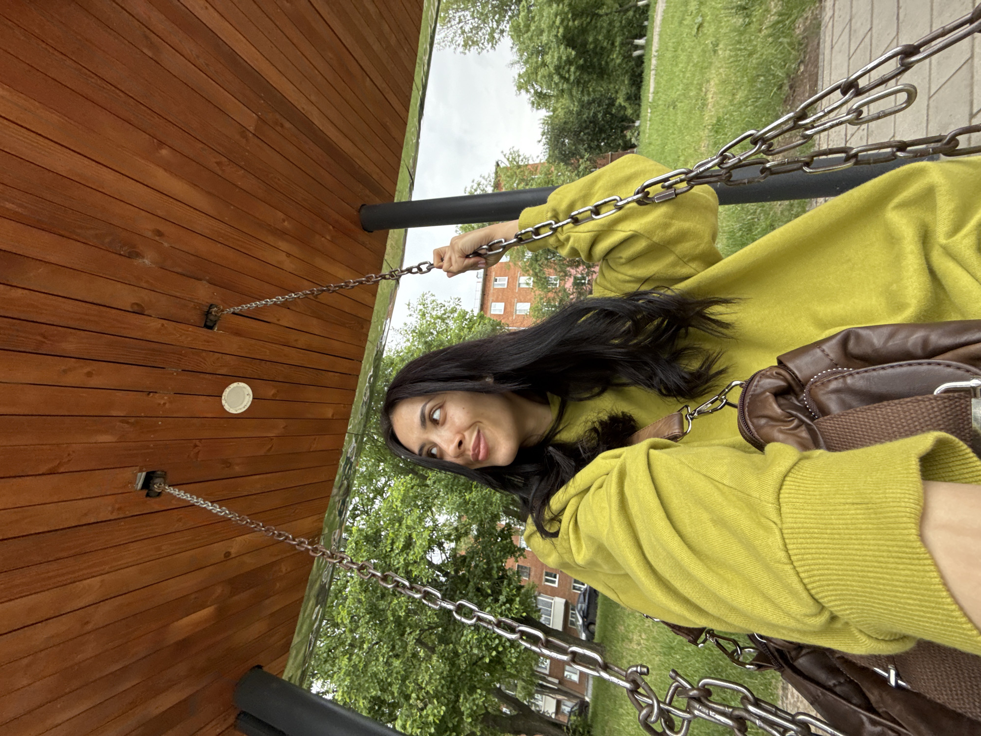 A young woman is captured in a close-up, half-body shot, sitting on a swing in what appears to be a park or playground setting in Votkinsk, Russia. She has long, dark, wavy hair and is wearing a lime green, long-sleeved top or sweatshirt with ribbed cuffs. A brown shoulder bag with silver hardware is visible on her right side. She holds onto the metal chains of the swing with her right hand and looks towards the viewer with a subtle, pleasant smile and a slightly upward gaze.
The swing is suspended from a sturdy wooden structure, featuring a slatted wooden ceiling. A small, white circular fixture, possibly a light or vent, is embedded in the wooden ceiling. A black cylindrical bar, likely part of the swing frame, is visible near her arm.
The background reveals lush green trees with dense foliage, suggesting a natural and verdant environment. Behind the trees, a multi-story residential building with reddish-brown walls and white window frames can be seen, indicating an urban or suburban area. A paved path or sidewalk is visible to the far right. The sky is overcast and cloudy, suggesting a daytime shot without direct sunlight. There is no visible text in the image. The overall impression is one of relaxed outdoor activity on a cloudy day.