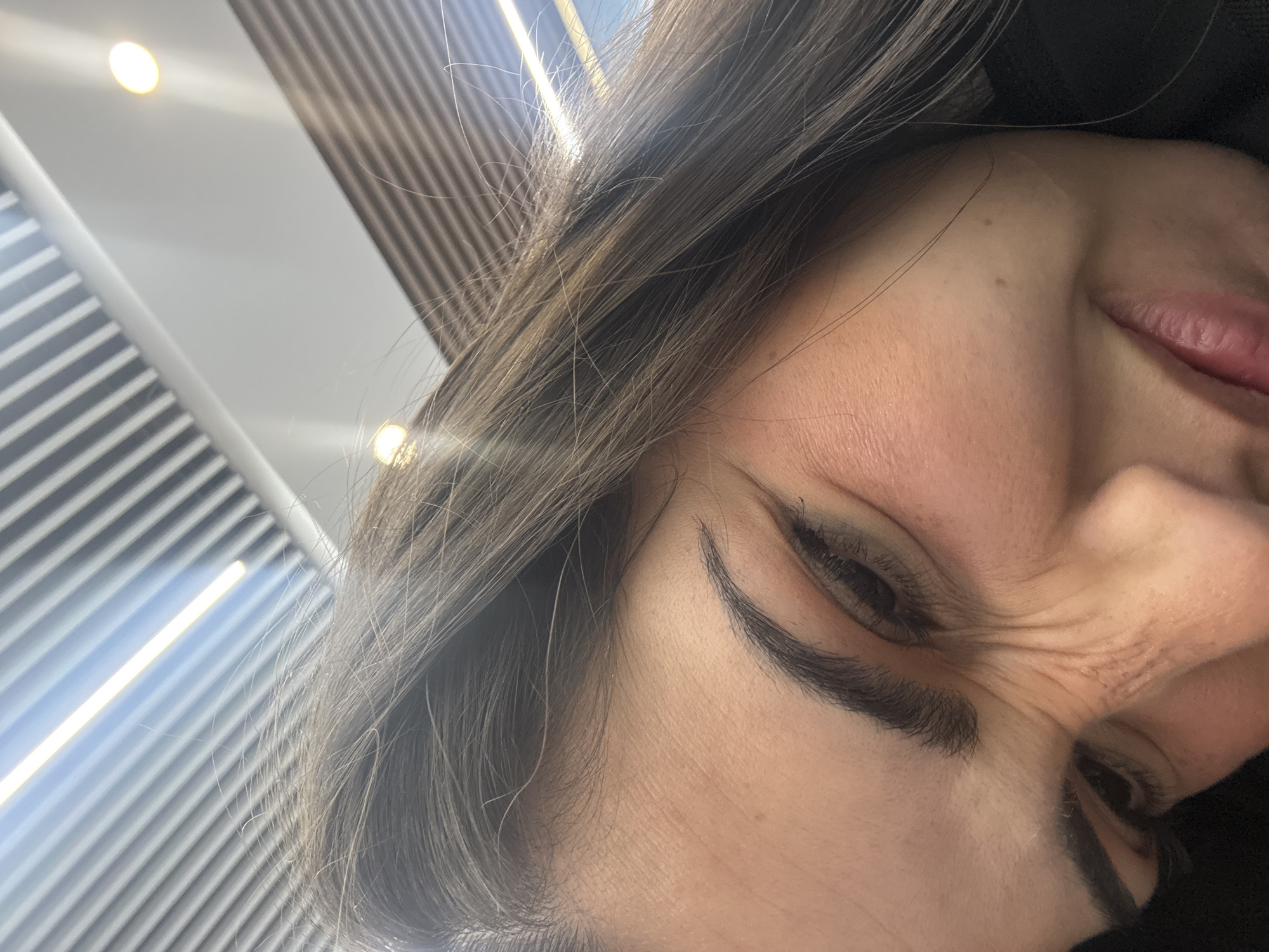 A close-up, angled view of a person with fair skin, brown hair, and dark eyebrows. The person's right eye is partially closed in a squint or smile. Wrinkles are visible around the eye and on the forehead. The person's lips are slightly parted, and the tip of their nose is visible. A black garment is visible behind the person's head. The background consists of a ceiling with linear architectural elements and visible light fixtures. The image appears to be taken indoors, possibly at a modern building in Mexico City, based on the architectural style and lighting.