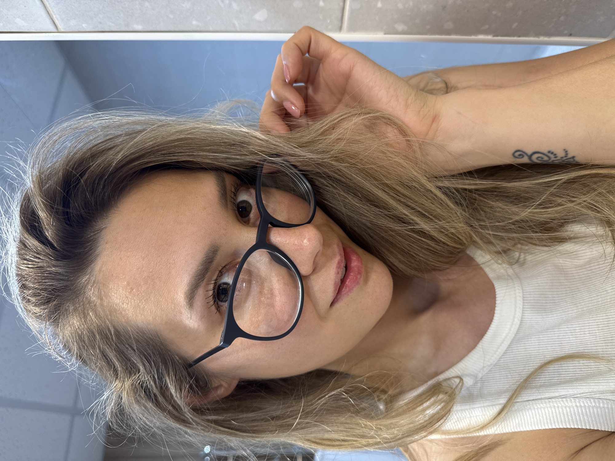 A young Caucasian woman with long blonde hair and glasses is pictured in a close-up selfie. She is wearing a white ribbed tank top. Her left hand is touching her hair, and a tattoo is visible on her left wrist. The background appears to be a bathroom with light blue walls and a tiled surface. The setting is indoors, likely a residential bathroom in the city of Nebug, Russia.