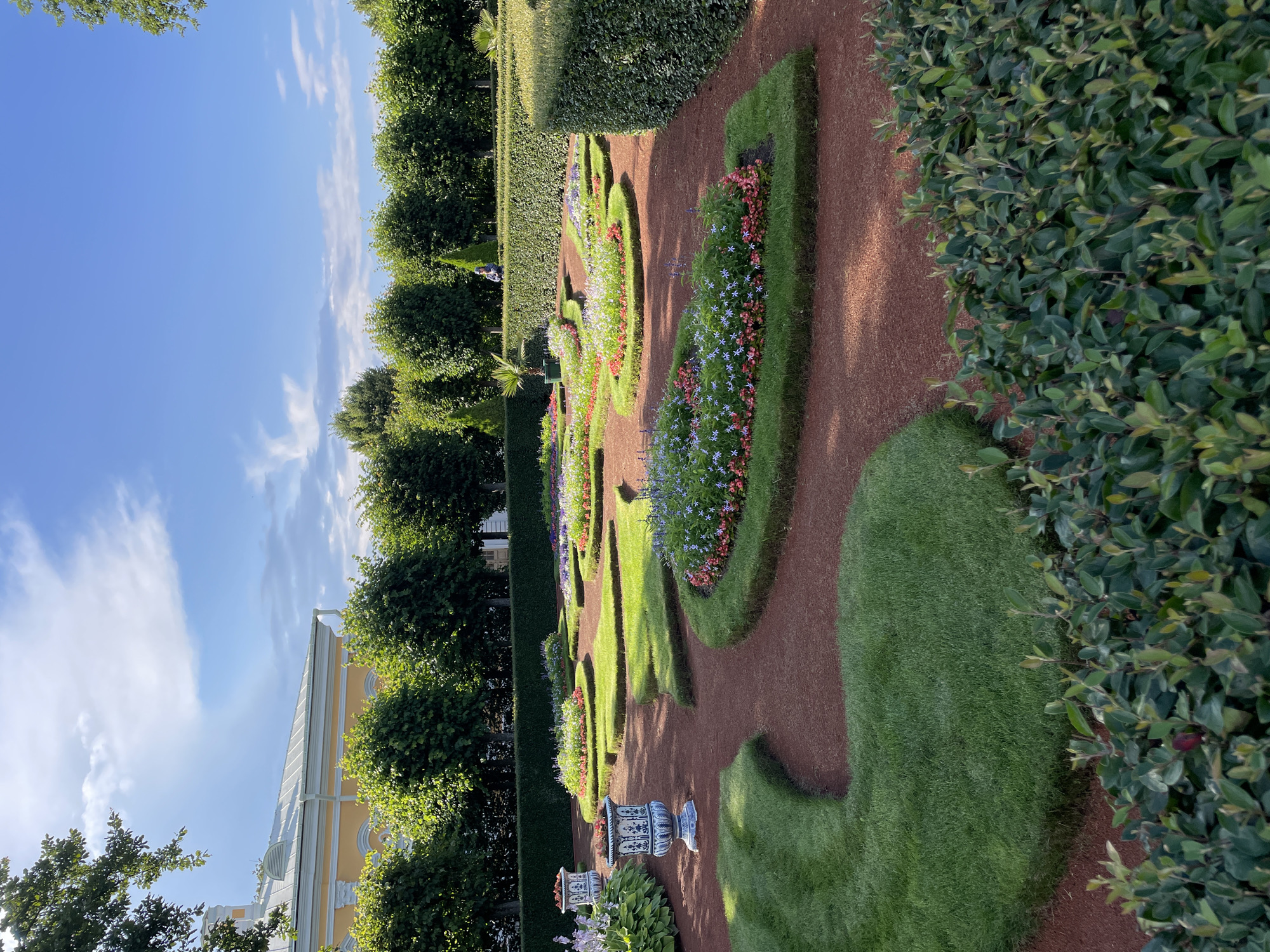 This image depicts a formal garden in the city of Peterhof, Russia. The garden features meticulously shaped green lawns interspersed with circular flowerbeds planted with colorful flowers. The pathways are covered in red gravel, and manicured hedges line the perimeter of the garden. In the background, a yellow building with a white roof is visible, along with a line of tall, dense trees. The sky is blue with scattered white clouds. One person is visible in the distance, standing on a pathway. The overall scene suggests a well-maintained public garden.