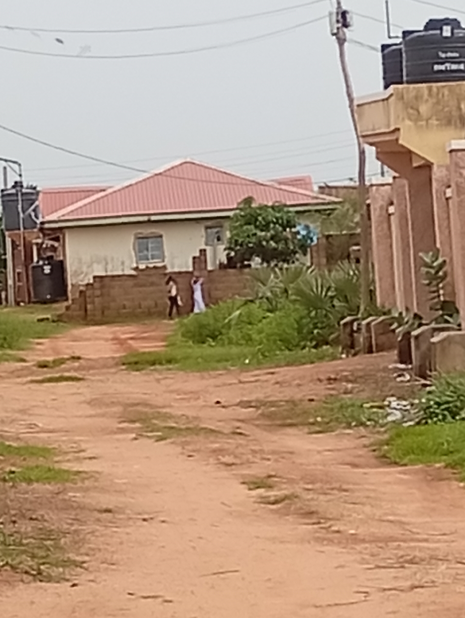 The image depicts a dirt road in a residential area of Tumfure, Nigeria. On the left side of the road, there is a single-story building with a red corrugated metal roof and beige walls. In front of the building, there is a low wall made of brick. Two individuals are visible walking near the building: one person is wearing a white garment, and the other is wearing a dark top and lighter pants. To the left of the building, a large black water tank is mounted on a stand. Further to the right, another building with a light brown facade and vertical brown pillars is visible. The environment is characterized by sparse vegetation and a cloudy sky. The ground is a reddish-brown dirt, indicative of a rural or semi-urban setting.