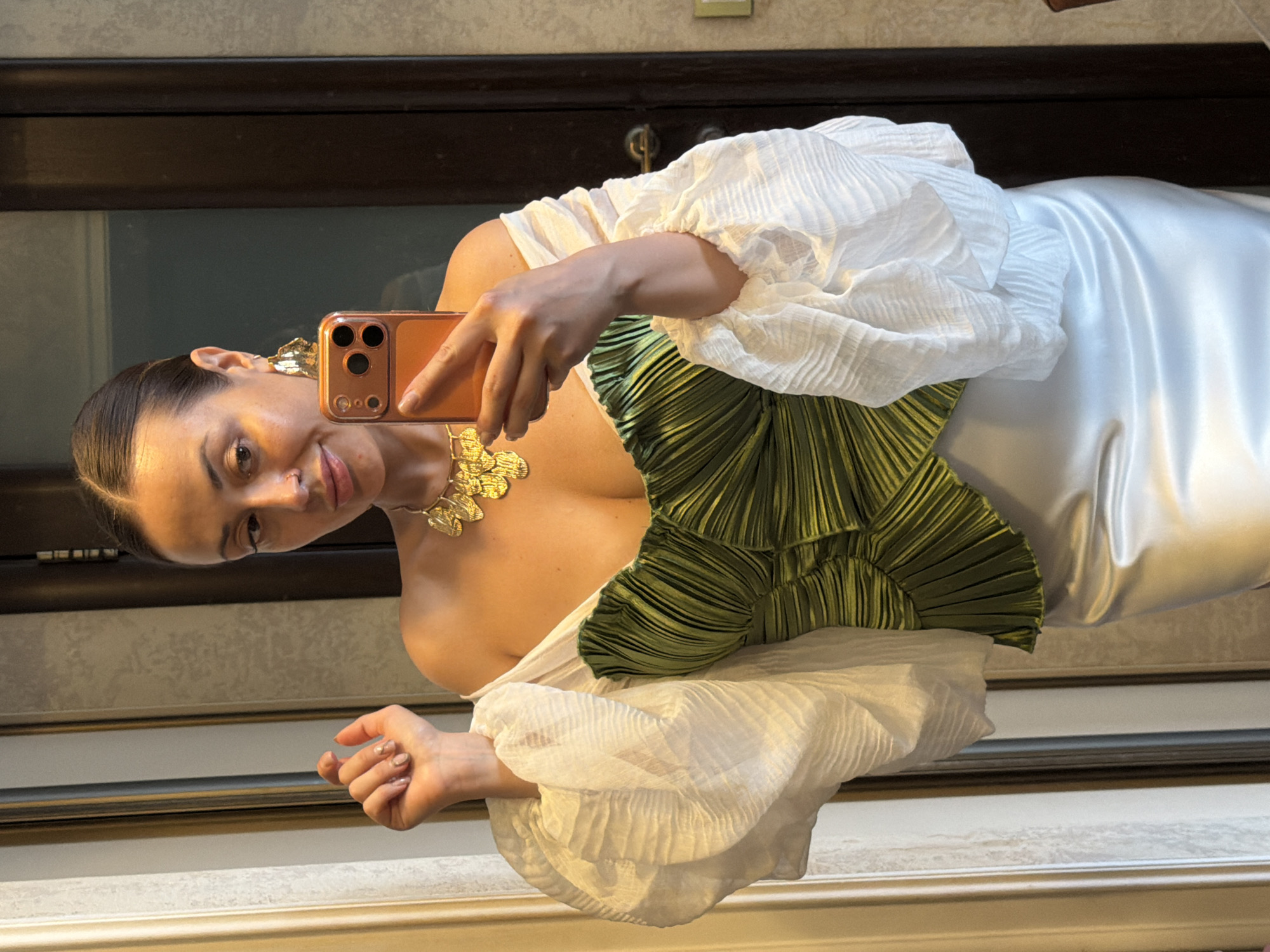 A woman with dark hair pulled back into a bun and light makeup is taking a mirror selfie. She is wearing a strapless, off-the-shoulder green pleated corset and a white sheer puff-sleeved top. She has a large gold statement necklace and large gold leaf-shaped earrings. She is holding a rose-gold colored smartphone in her right hand to take the picture. The background appears to be an indoor setting, possibly a home, with a reflective surface, likely a mirror, capturing her image. There is a dark wooden door or frame visible in the background, and part of a wall. The lighting is warm and casts shadows. The overall scene is a personal portrait captured through a reflection. The image does not contain any elements that can identify the location as Krasnoyarsk, Russia.